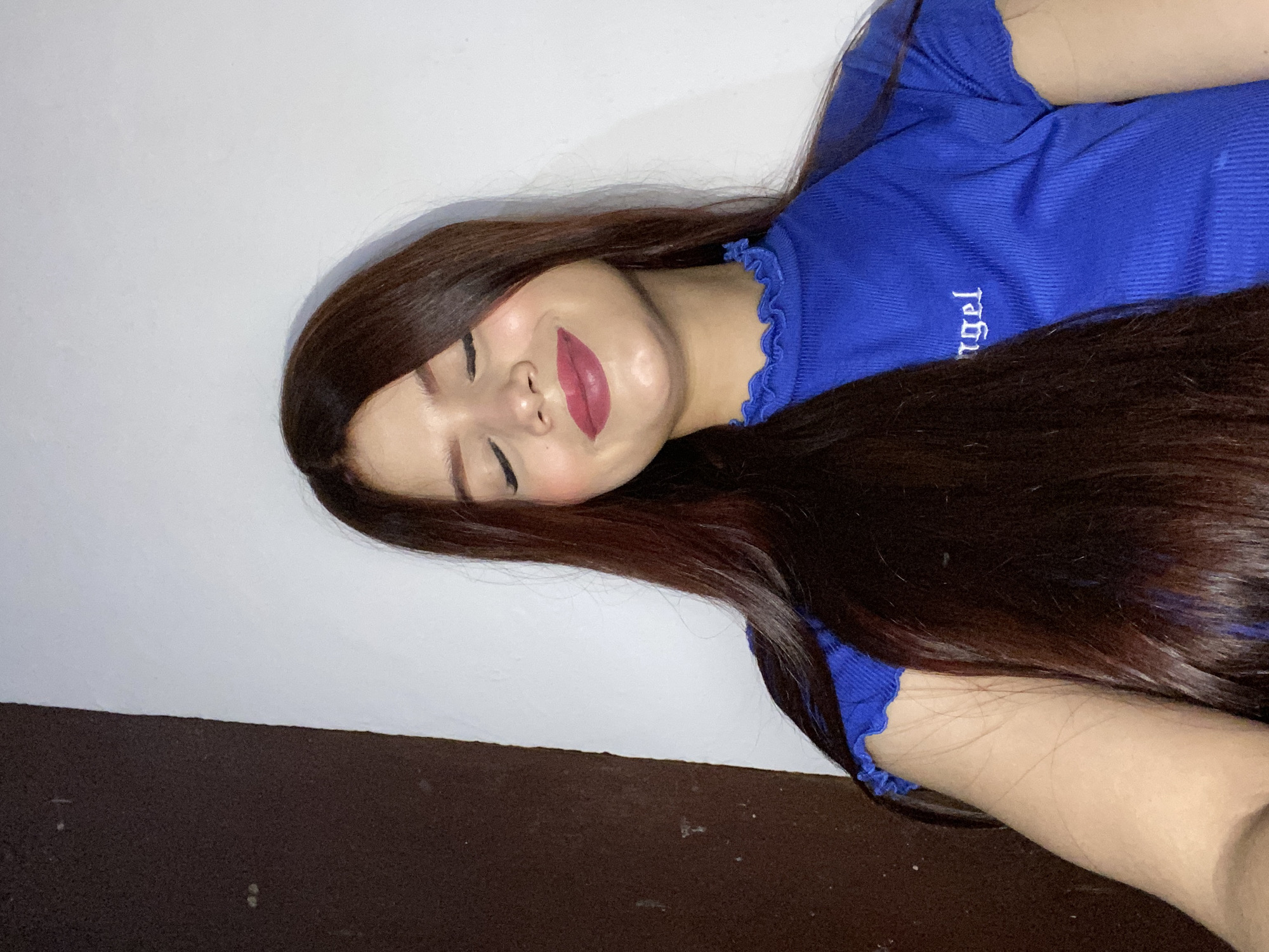 A young woman is lying on her side against a white wall above a dark brown surface. She has long, dark brown hair and is wearing a blue top with the word "angel" vertically printed on it. Her eyes are closed, and she is wearing dark pink lipstick. The photograph appears to be taken indoors. I don't see any elements that indicate it was taken in Juan Griego, Venezuela.
