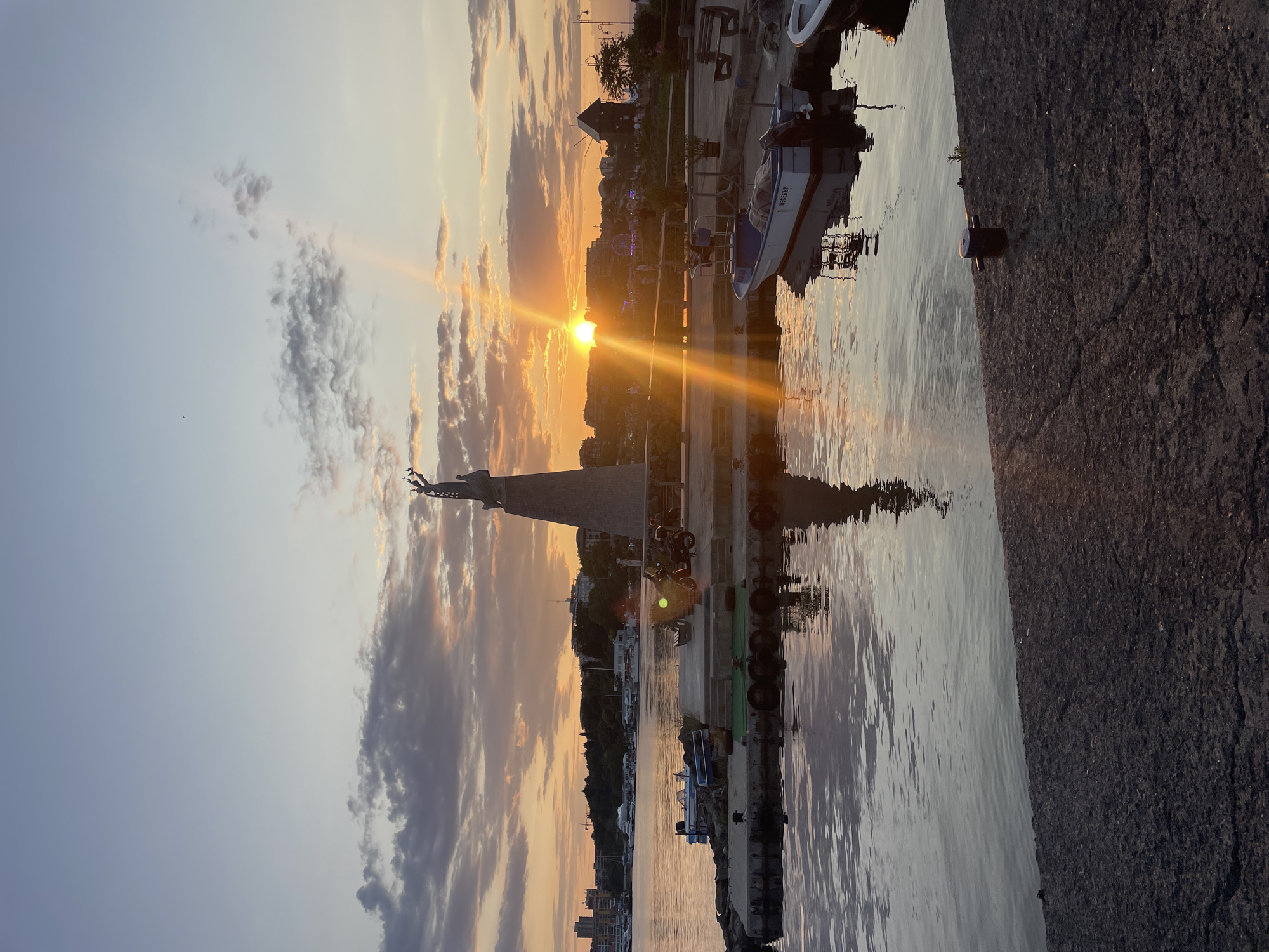 The image depicts a sunset over the Black Sea in Nesebar, Bulgaria. A large statue of a woman holding a cross stands prominently on a concrete platform by the water. The sun is setting behind a distant cityscape, casting a golden glow across the sky and reflecting on the water. Several boats are docked in the harbor, with their reflections visible on the calm surface of the sea. A windmill is visible in the background, adding to the scenic coastal atmosphere. The foreground shows a textured concrete walkway leading to the water. The sky is filled with scattered clouds tinged with orange and pink hues from the setting sun.