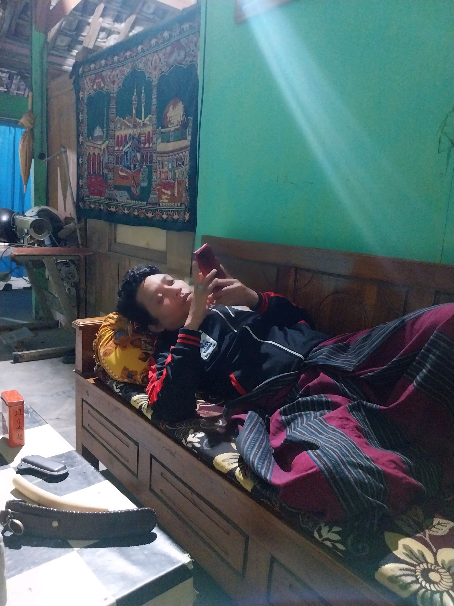 A person is lying on a wooden bench, looking at a mobile phone. The person is wearing a black and red jacket and a sarong. The sarong is red with dark blue and white stripes. The bench is covered with a patterned fabric and a yellow pillow. In the background, there is a green wall with a tapestry hanging on it. The tapestry depicts a mosque and minarets. To the left of the tapestry, there is a blue curtain and a sewing machine. On the table in the foreground, there are some objects, including a red box with Chinese characters, a black leather case, and a brown leather sheath with a knife. The room appears to be a living space in a house in Campur, Indonesia.