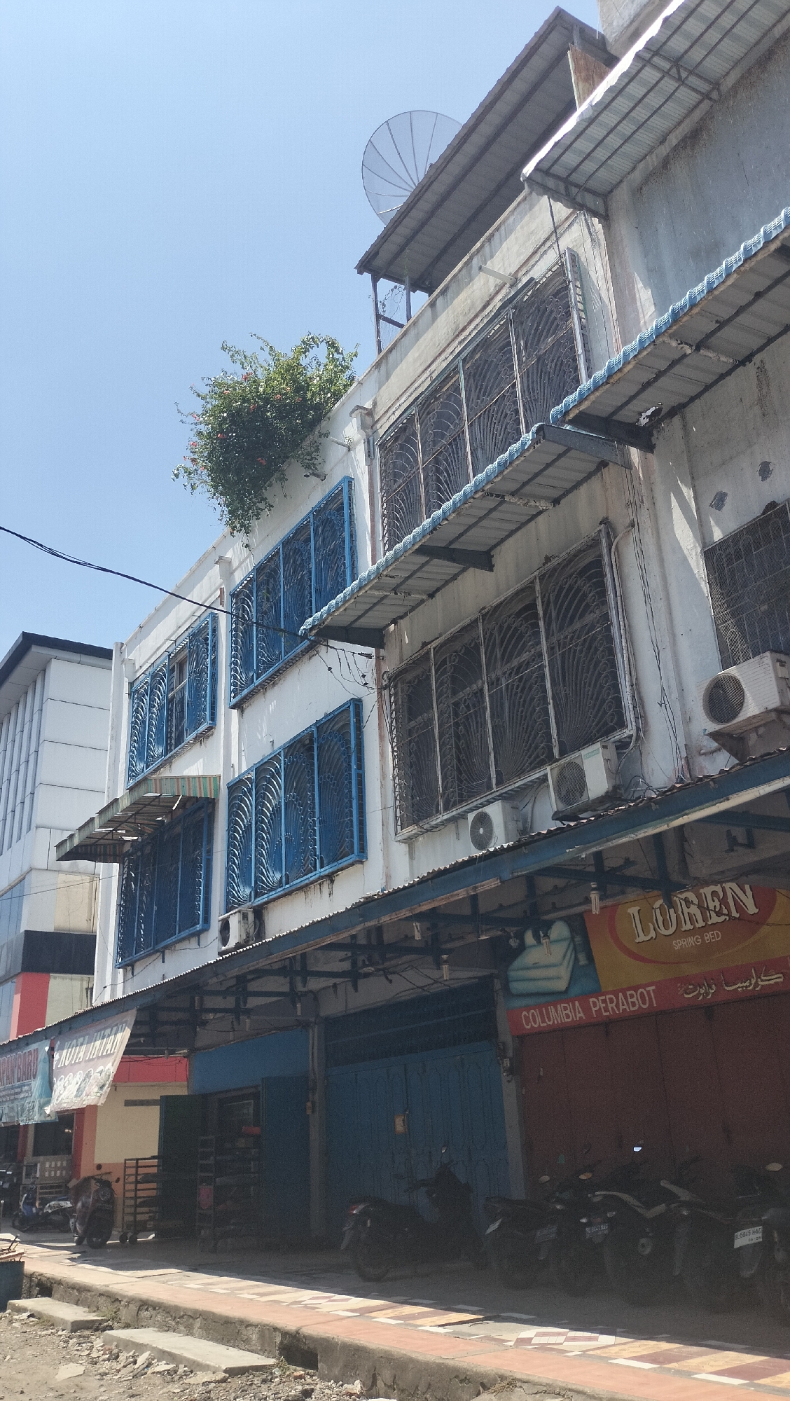 The image depicts a street scene in Lhokseumawe, Indonesia, on a bright, sunny day. The focus is on a multi-story building with a white facade, featuring numerous windows covered by decorative blue metal grilles. The upper floors have awnings extending outwards, and air conditioning units are visible. Lush green foliage with some red flowers cascades from the top of the building. To the left, another building with vertical white panels is partially visible. The ground floor of the main building features shops with metal shutters and a prominent sign that reads "LOREN SPRING BED" in English and another sign in Arabic script. Several motorcycles are parked along the sidewalk in front of the shops. The street in the foreground appears to be paved, with a concrete curb and some debris. The sky is clear and blue.