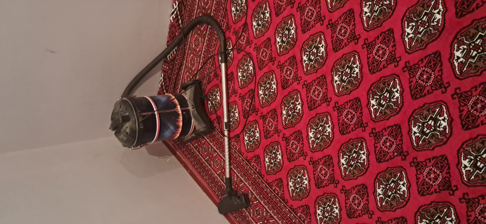A black canister vacuum cleaner with a decorative band of flames sits on a red rug with a repeating geometric pattern. The vacuum has a dark gray base and a black top with a handle. A black hose extends from the top of the vacuum and is connected to a metal wand with a black vacuum head lying on the rug. The rug has a border with a triangular pattern. The scene is indoors, likely in a room with a plain gray wall in the background. The image does not provide any specific information about the location in Yazy, Turkmenistan.