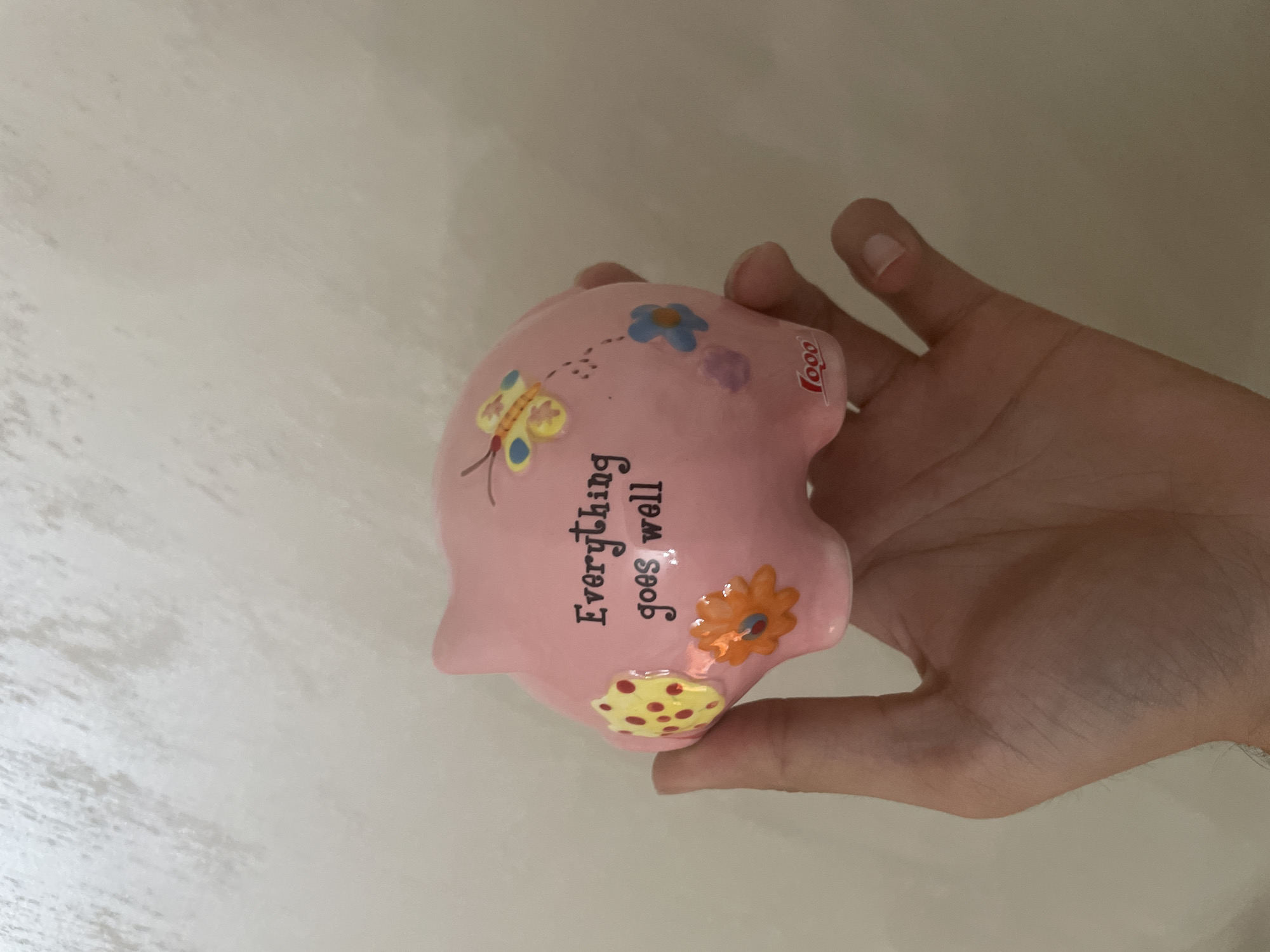 A human hand, appearing to be an adult's right hand with visible veins and short nails, holds a pink ceramic piggy bank against a light-colored, subtly textured wall. The piggy bank is adorned with various colorful decals, including a yellow butterfly with blue wings, an orange flower, a blue flower, and a yellow polka-dotted patch. The phrase "Everything goes well" is written in black script on the side of the piggy bank. A small red logo, appearing to read "Logo," is visible near the base of the piggy bank. The scene is situated in the city of Ihtiman, Bulgaria. No other individuals or objects are present, and the lighting is even and diffused.