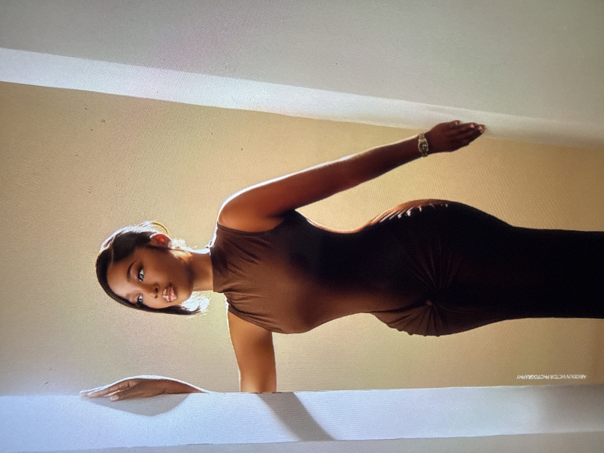 A woman is posing on a white surface, her body positioned in a plank-like stance. She is wearing a brown, sleeveless, form-fitting dress. Her left arm is extended forward, with her hand resting on the white surface. Her right arm is bent and raised behind her head, and she is wearing a watch on her right wrist. Her head is tilted and turned towards the viewer, with her eyes looking directly at the camera. Her hair is pulled back. The background consists of a light brown wall and a white ceiling, with a distinct architectural molding where the wall meets the ceiling. The lighting suggests it might be daytime, with light coming from above and to the side, casting a shadow. A faint watermark reading "ABDULN VICTOR PHOTOGRAPHY" is visible in the bottom right corner of the image. The image appears to be taken indoors, likely in a studio or a well-lit room, given the controlled lighting and clean background. The woman's pose and gaze suggest a professional photoshoot. The overall tone is elegant and focused. The location context is identified as Abuja, Nigeria.