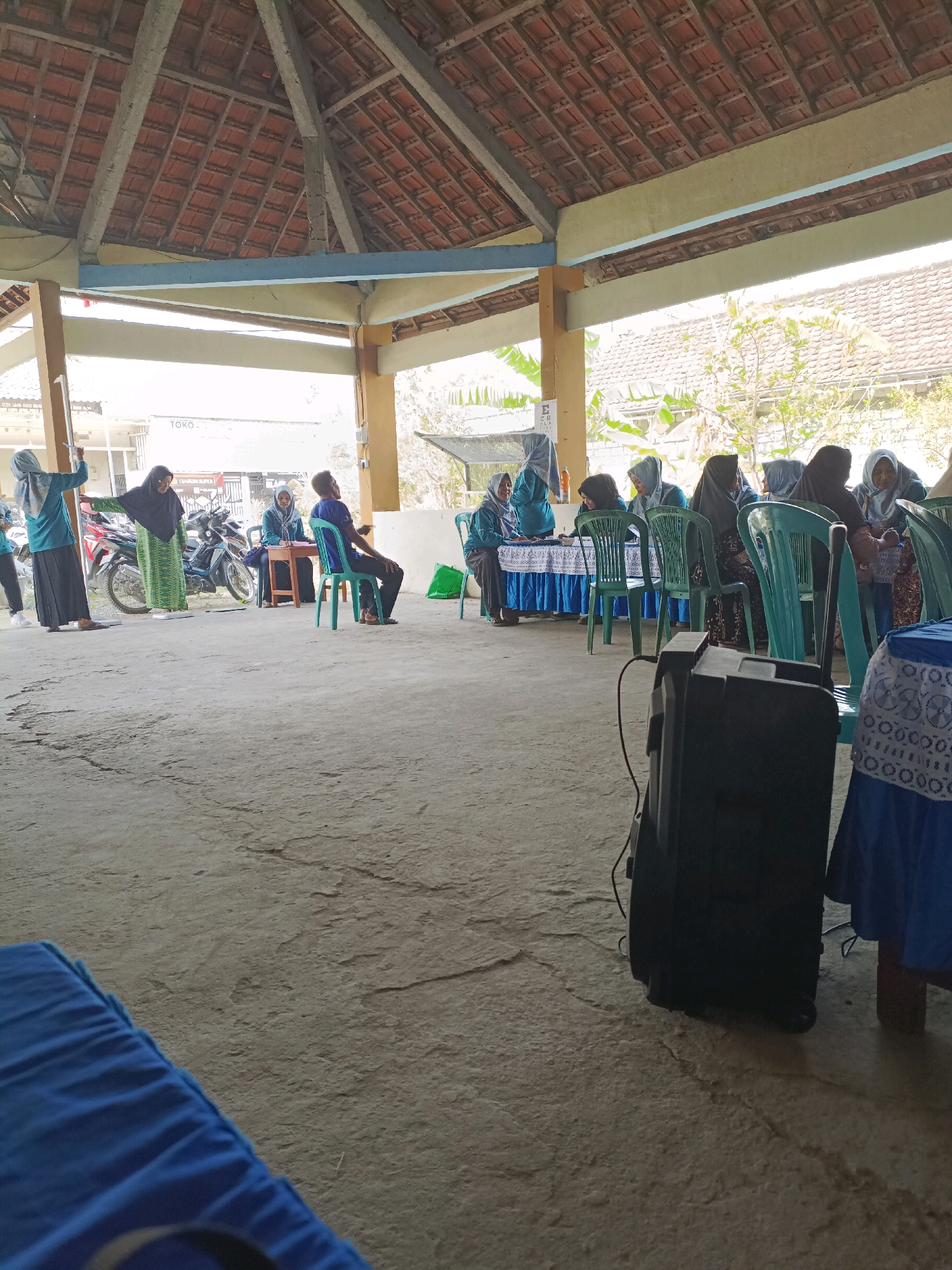 In an open-air pavilion with a terracotta-tiled roof supported by wooden beams, a group of people, predominantly women wearing hijabs, are gathered. The scene appears to be a community event or meeting in Rancang Kencono, Indonesia. In the foreground, to the left, a blue tarp is visible, and to the right, a black speaker stands on the concrete ground.
Several individuals are seated at tables draped with blue and white tablecloths, some appearing to engage in registration or information dissemination. A woman in a green patterned dress and black hijab stands near a motorcycle, seemingly directing attention towards something. Another woman in a teal hijab and black skirt stands beside her, holding a stick or pole. A man in a blue shirt and black pants is seated in a green chair, facing away from the camera. Further back, more individuals are seated at tables, engaged in conversation or activities. Motorcycles are parked in the background, suggesting a mode of transportation for attendees. The environment is a concrete floor with visible cracks, and beyond the pavilion, glimpses of buildings and trees suggest an outdoor setting within a populated area.