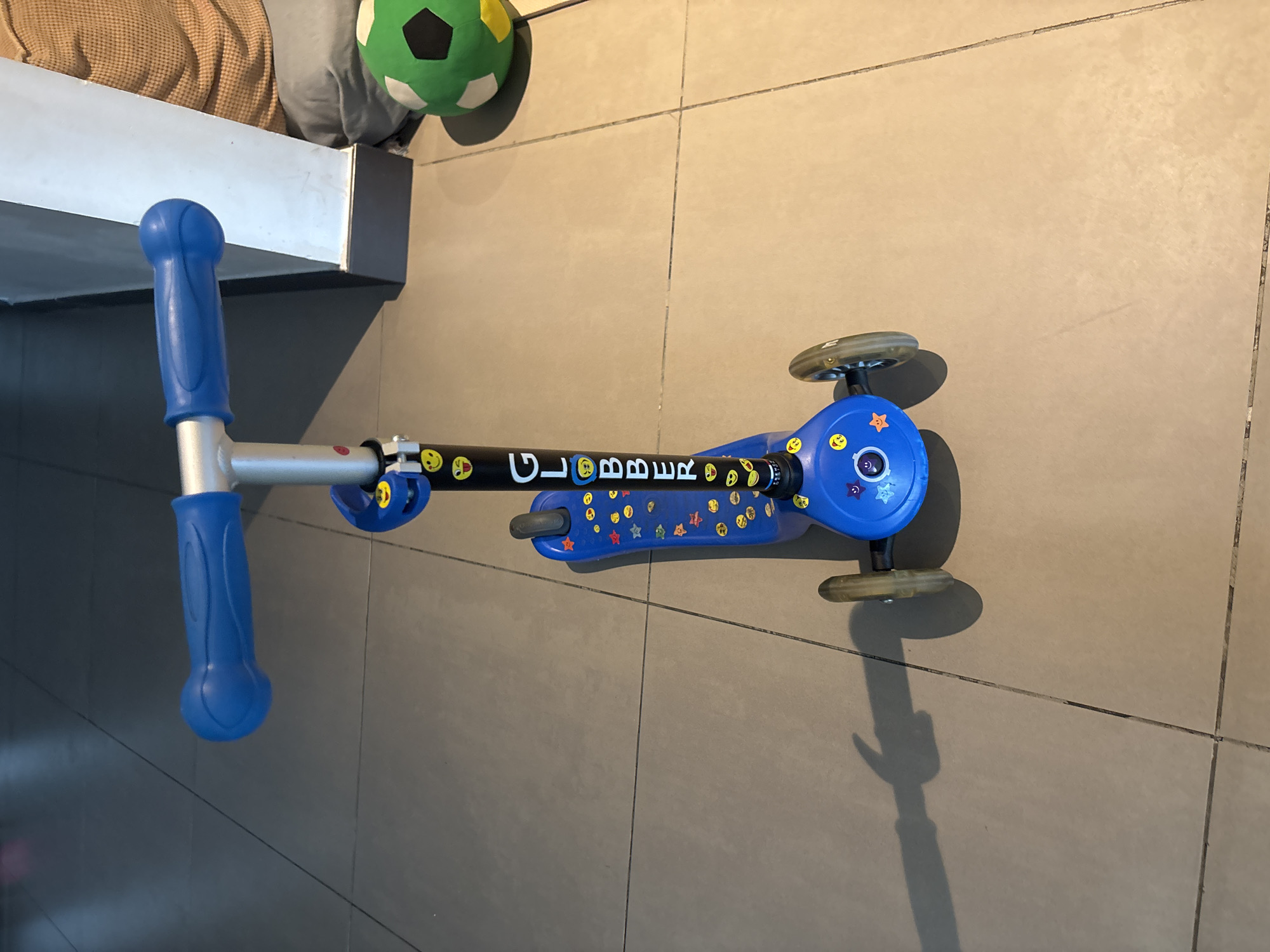 A blue three-wheeled scooter with smiley face stickers and the brand name "GLOBBER" written vertically on the handlebar stem is positioned on a tiled floor. The scooter has blue handlebar grips and a blue deck. To the right of the scooter, a green and white soccer ball is visible. The background appears to be an indoor setting with a wall and a glimpse of a person wearing a tan shirt. The lighting suggests an indoor environment. There is no indication of the specific location within Dubai, United Arab Emirates.