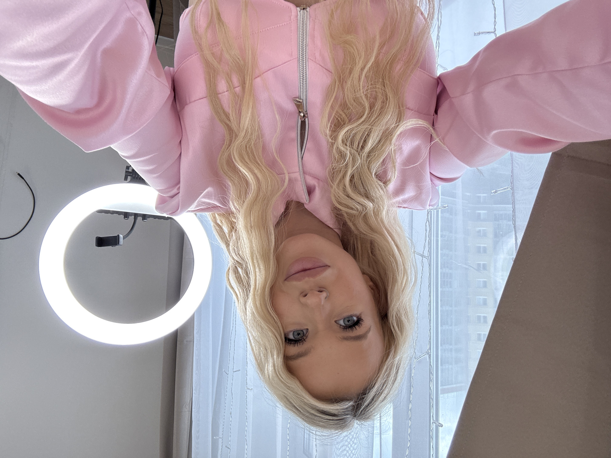 The image is an upside-down, close-up selfie of a young woman with blonde, wavy hair. She is wearing a light pink hooded jacket with a zipper. Her eyes are blue and she is looking directly at the camera with a neutral expression. She has a small nose ring.
To her left, a circular, bright LED ring light is visible, attached to a black mount. Behind her, sheer white curtains are drawn, and through them, a blurry image of a modern apartment building can be seen, suggesting an urban setting, potentially in Saint Petersburg, Russia, as indicated by the location context. Decorative string lights are draped along the window frame. The overall lighting suggests it might be daytime. The context of the ring light and selfie suggests the woman may be creating content or engaging in online communication.