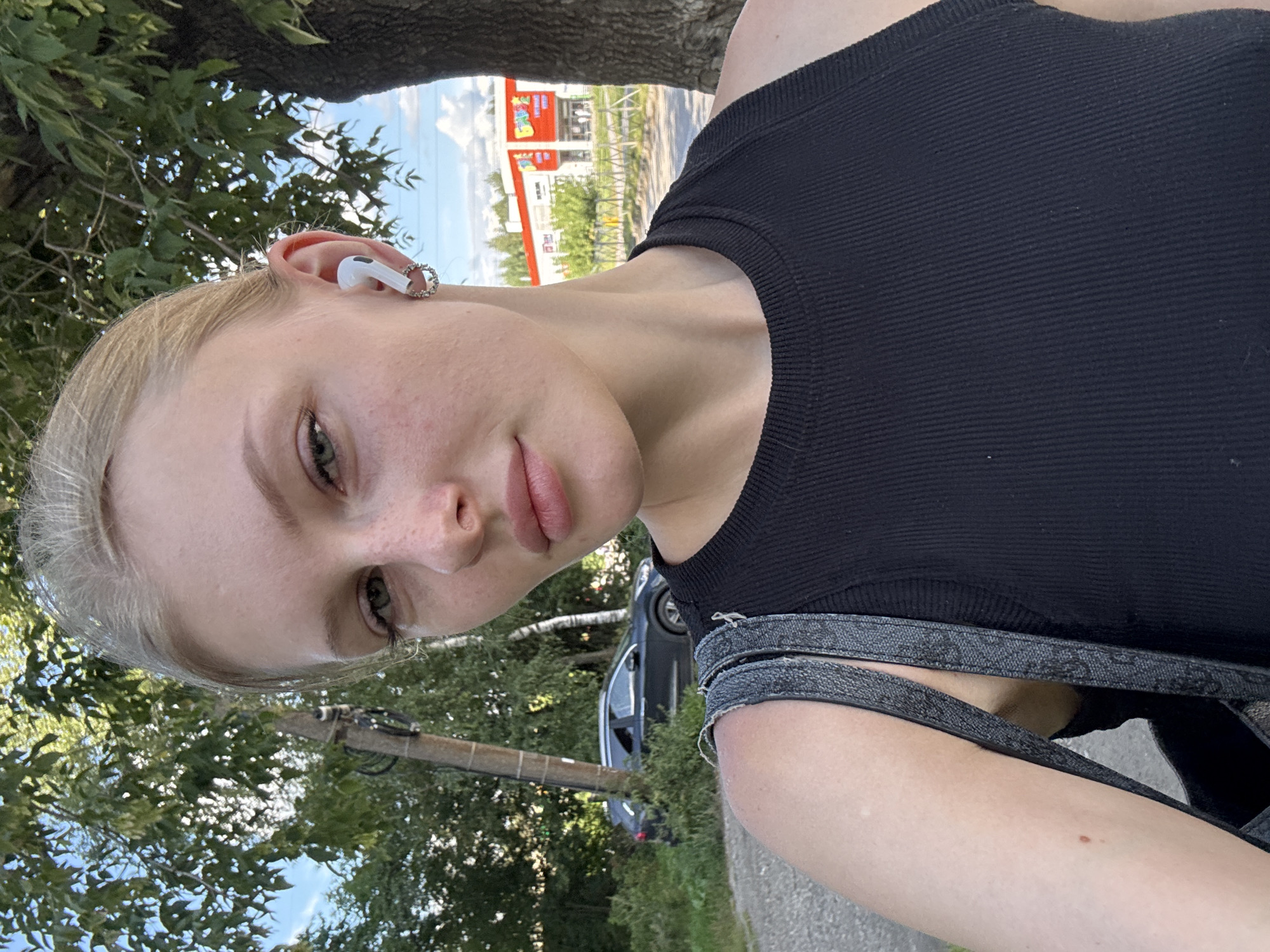 A young woman with blonde hair pulled back, wearing a black ribbed tank top and white earbuds, takes a selfie outdoors. She has fair skin with freckles and is wearing a small hoop earring on her visible ear. Behind her, trees and a dark-colored SUV parked on the side of a road are visible. In the background, a red building with signage can be seen, possibly a commercial establishment. The overall lighting suggests it is daytime. The presence of trees, a road, and a commercial building indicates an urban or suburban setting, consistent with a city location like Perm, Russia.