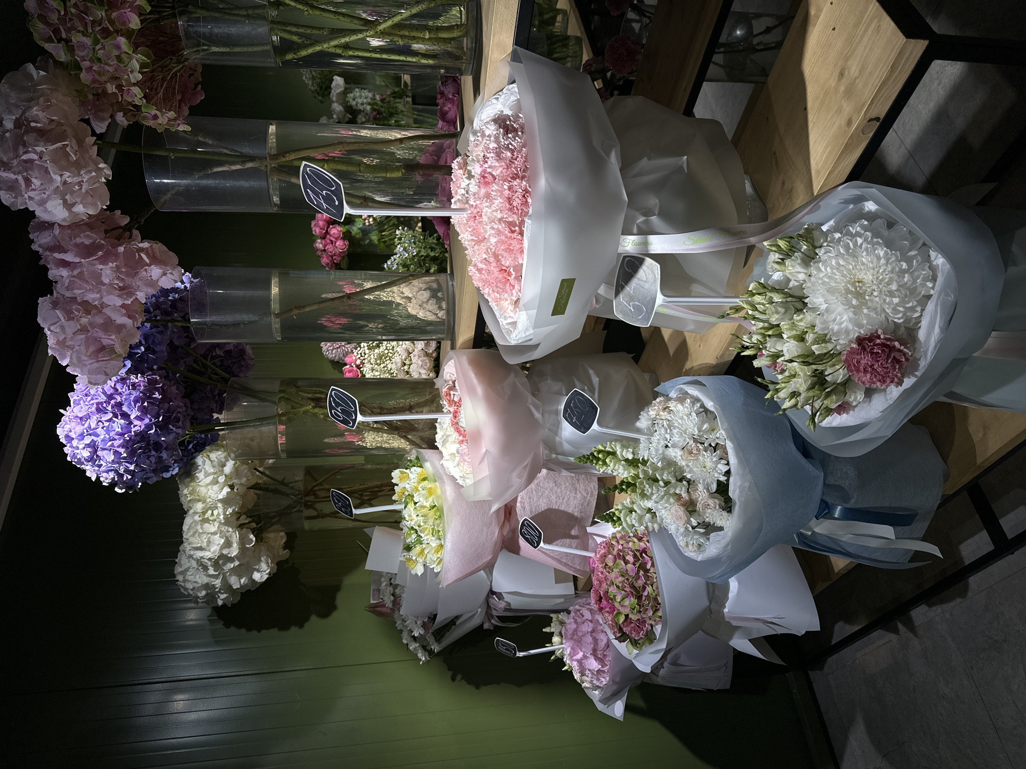 The image shows a display of flowers in what appears to be a flower shop or market, possibly in Chișinău, Moldova, although location confirmation is not explicitly available. The flowers are arranged on a tiered structure, likely a shelf or stand. On the left, there are various types of flowers, including hydrangeas in shades of pink, purple, and white, arranged in clear glass cylindrical vases filled with water. Each vase contains a single type of flower. Small, black rectangular price tags are stuck into the vases. To the right, there are several wrapped bouquets of different floral arrangements. The bouquets are wrapped in white and pink paper with ribbon accents. Price tags are also visible on these bouquets. The lighting in the image appears to be a mix of natural and artificial light, casting shadows and highlights on the flowers and their packaging. The overall environment seems clean and organized, suggesting a well-maintained retail space. The angle of the photograph gives a slightly elevated perspective of the flower display. A wooden structure forms a part of the shelves containing the flower bouquets. The bottom of the image shows the floor and some wall space.