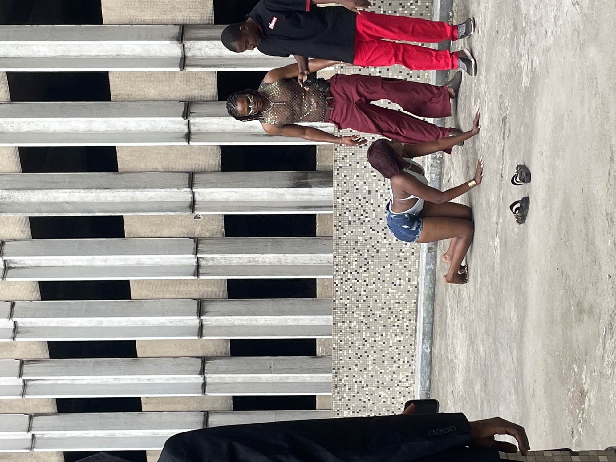 The image shows a group of four people posing, likely for a photoshoot, in an urban environment, possibly in Lagos, Nigeria.
One person, wearing a black shirt and red pants, stands at the top of the image, looking down. Another person is wearing a mesh top and maroon pants, posing with arms extended. A third person is kneeling with a white tank top and denim shorts. Nearby are a pair of sandals. In the foreground, a portion of a person's arm and jacket is visible, holding a phone.
The background includes a wall with a stacked pattern of white and black blocks, along with a tiled surface composed of small, square tiles. The area appears to be a rooftop or open-air location.