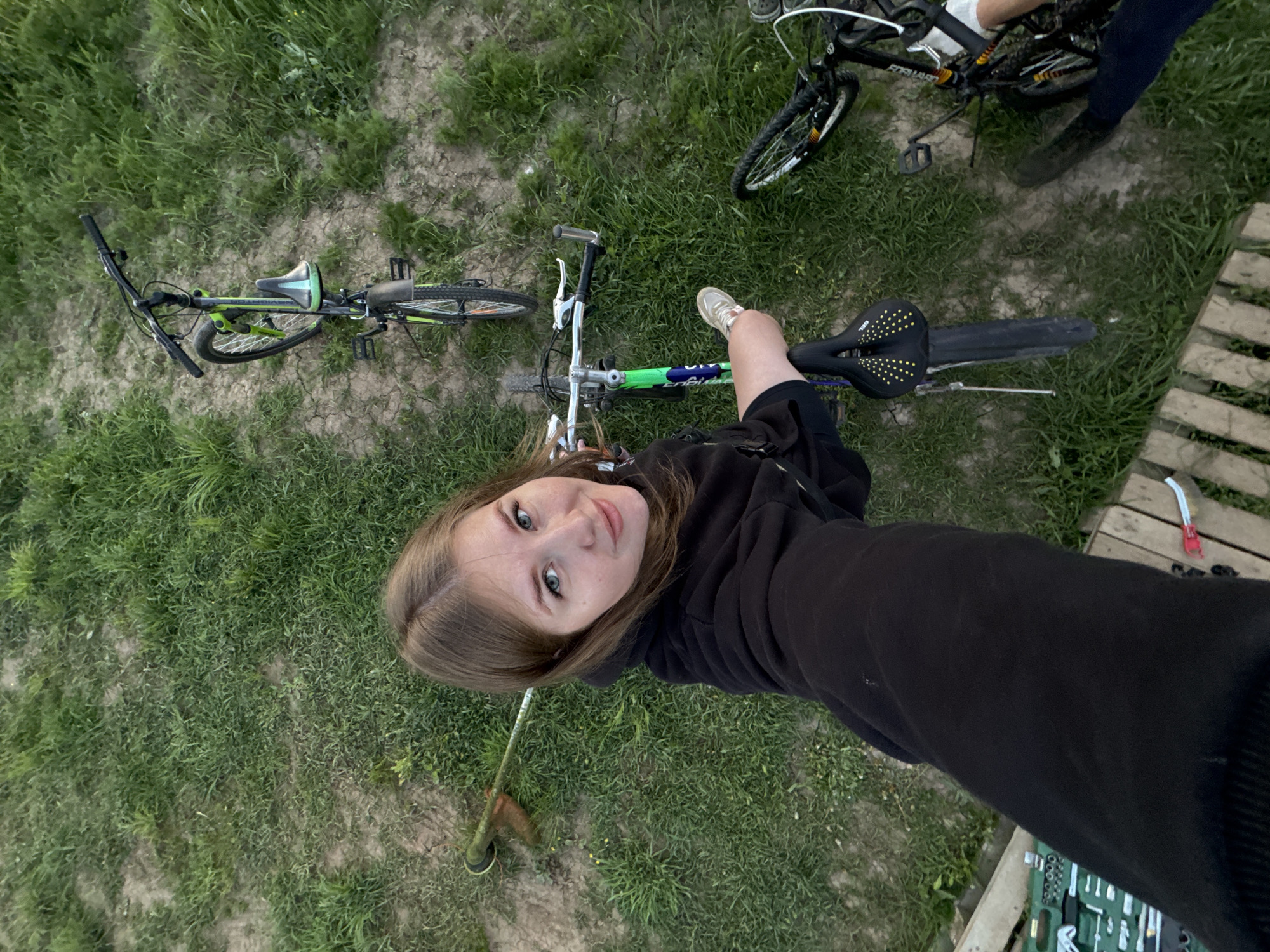 The image, taken from an elevated perspective, depicts a woman in a black hooded sweatshirt standing on a wooden pallet amidst a grassy area in what appears to be an outdoor setting. A green and blue bicycle lies horizontally across the frame, partially overlapping the woman's leg. To the upper right, another bicycle is visible, partially cropped. A string trimmer rests on the grass near the woman. To the bottom right, a tool kit is partially visible on the wooden pallet. The location is ферма, Russia.
