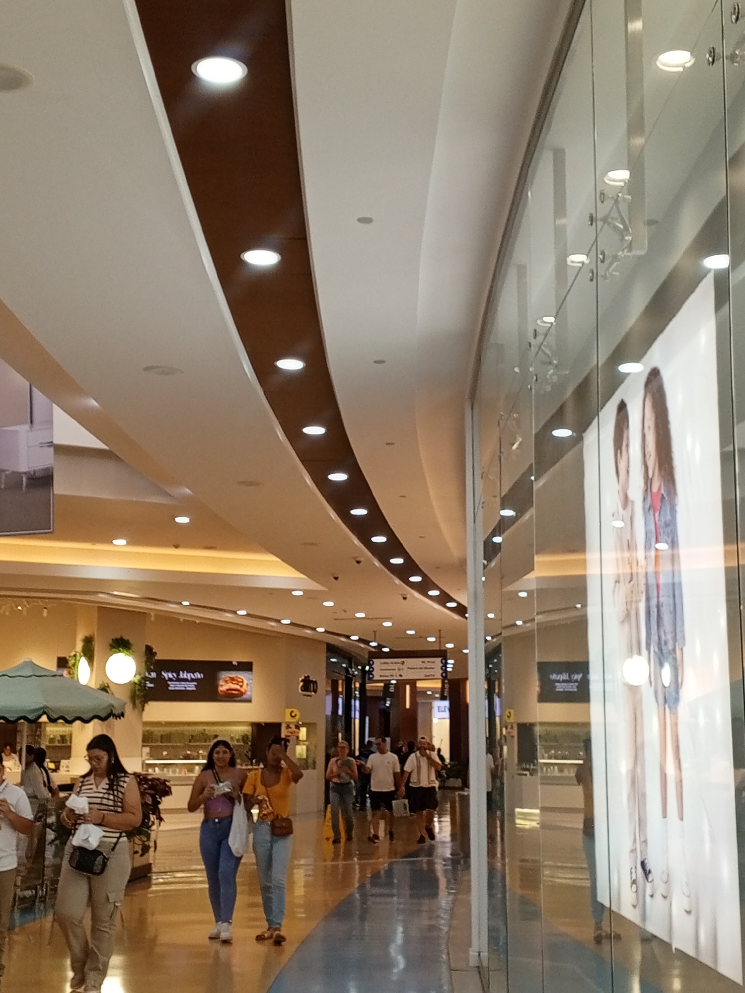 The photograph captures a scene inside a shopping mall. The interior features curved architectural elements, including a brown strip running along the ceiling with evenly spaced recessed lighting. The floor is polished and reflective. On the right side, a storefront with a curved glass facade displays a large advertisement featuring images of women. People are walking along the mall corridor, including a group of three women walking toward the camera. The background contains various shops and eateries, identifiable by their signage. A green canopy of a food stand is visible on the left side. The scene appears well-lit, with a mix of natural and artificial light sources. The image is taken in Porlamar, Venezuela.