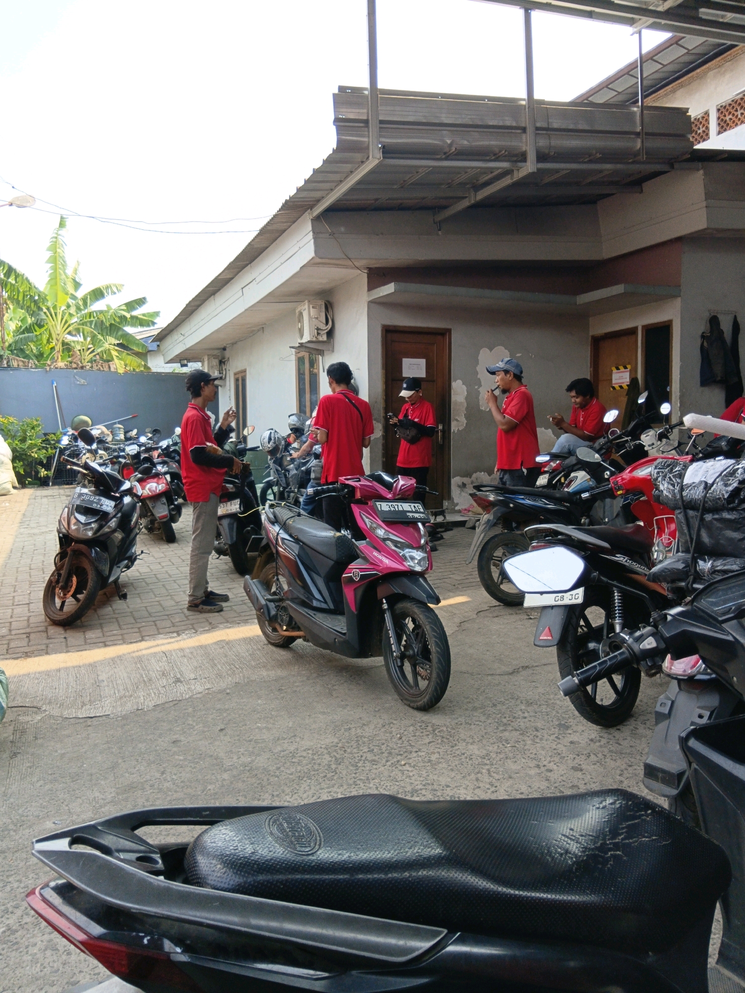 The image depicts a group of men in red shirts, likely delivery drivers, standing around several parked motorcycles in what appears to be a depot or storage area in Bekasi, Indonesia. The environment is an outdoor paved area with a low-rise building in the background. The building has a corrugated metal roof extension and several windows and doors. The men are engaged in various activities: some are looking at their phones, one is smoking and holding a phone, and another is talking. The motorcycles are a mix of scooter-style vehicles, common for delivery services. The overall atmosphere suggests a moment of pause or preparation before or after delivery runs. The presence of an air conditioning unit on the building and lush green vegetation in the background hint at a tropical or subtropical climate.