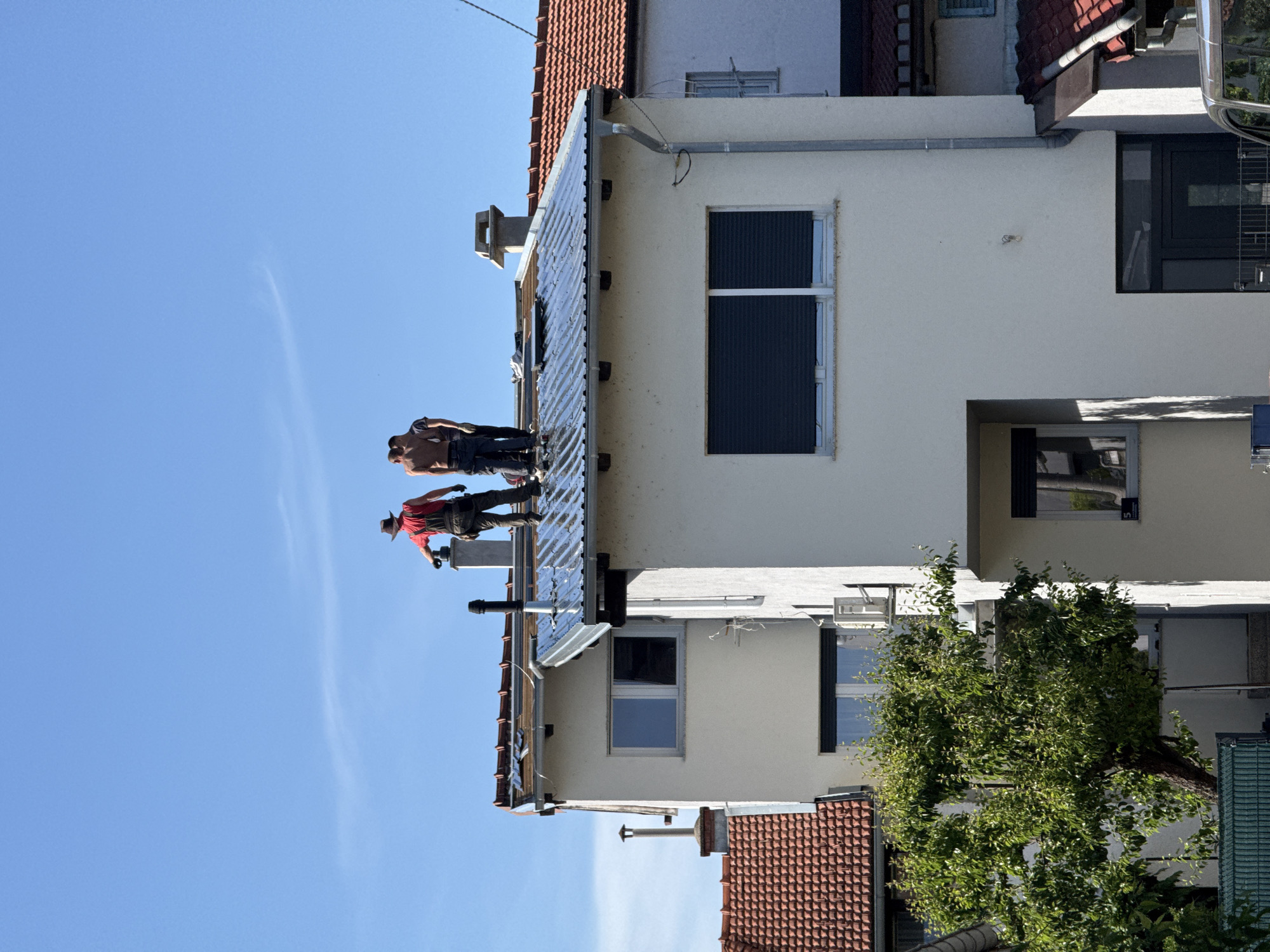 Two men are on the roof of a building. One is shirtless and the other wears a red shirt and hat. The roof is corrugated metal. The building is white with red roof tiles. There are windows on the building. The sky is blue. The image may be in City of Zagreb, Croatia, but there is no way to confirm this from the image alone.
