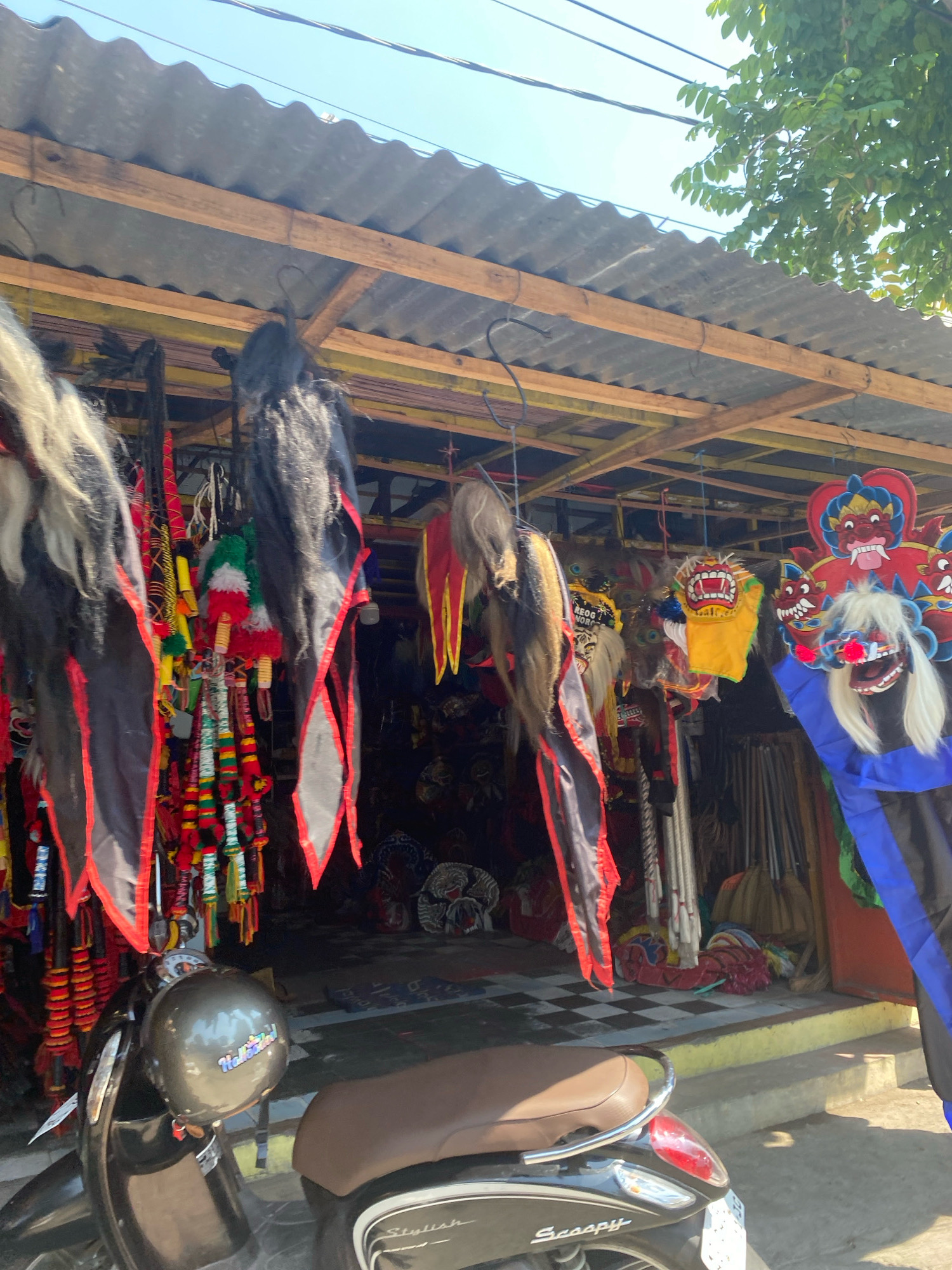 This is an eye-level, outdoor shot of a roadside stall in Kediri City, Indonesia. The stall is displaying traditional Indonesian folk art, likely for sale. The foreground shows a dark-colored scooter with a brown seat. Hanging from the stall's wooden frame are numerous colorful masks and decorative items. These include elaborately designed masks with striking features and long, flowing hair made of yarn or fabric, as well as bundles of colorful tassels and ornaments. The stall's roof is made of corrugated metal. The background reveals more of the stall's interior with stacked items and what appear to be poles or staffs. The ground in front of the stall has a checkered pattern. The scene is lit by bright sunlight, casting shadows, and a clear blue sky is visible. The overall impression is of a vibrant cultural marketplace.