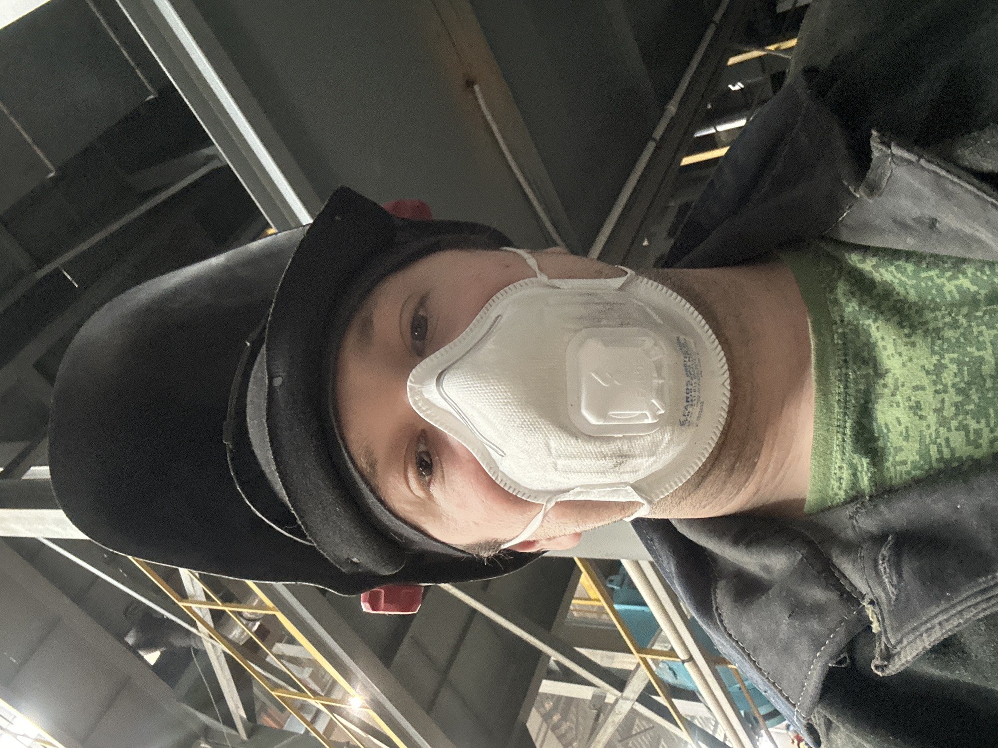 A person wearing a black welding helmet and a white particulate respirator is looking at the camera. The person also wears a dark jacket and a green patterned shirt. The background consists of metallic structures and beams, painted yellow and gray. The specific location within Noviy, Russia is not discernible from the image.