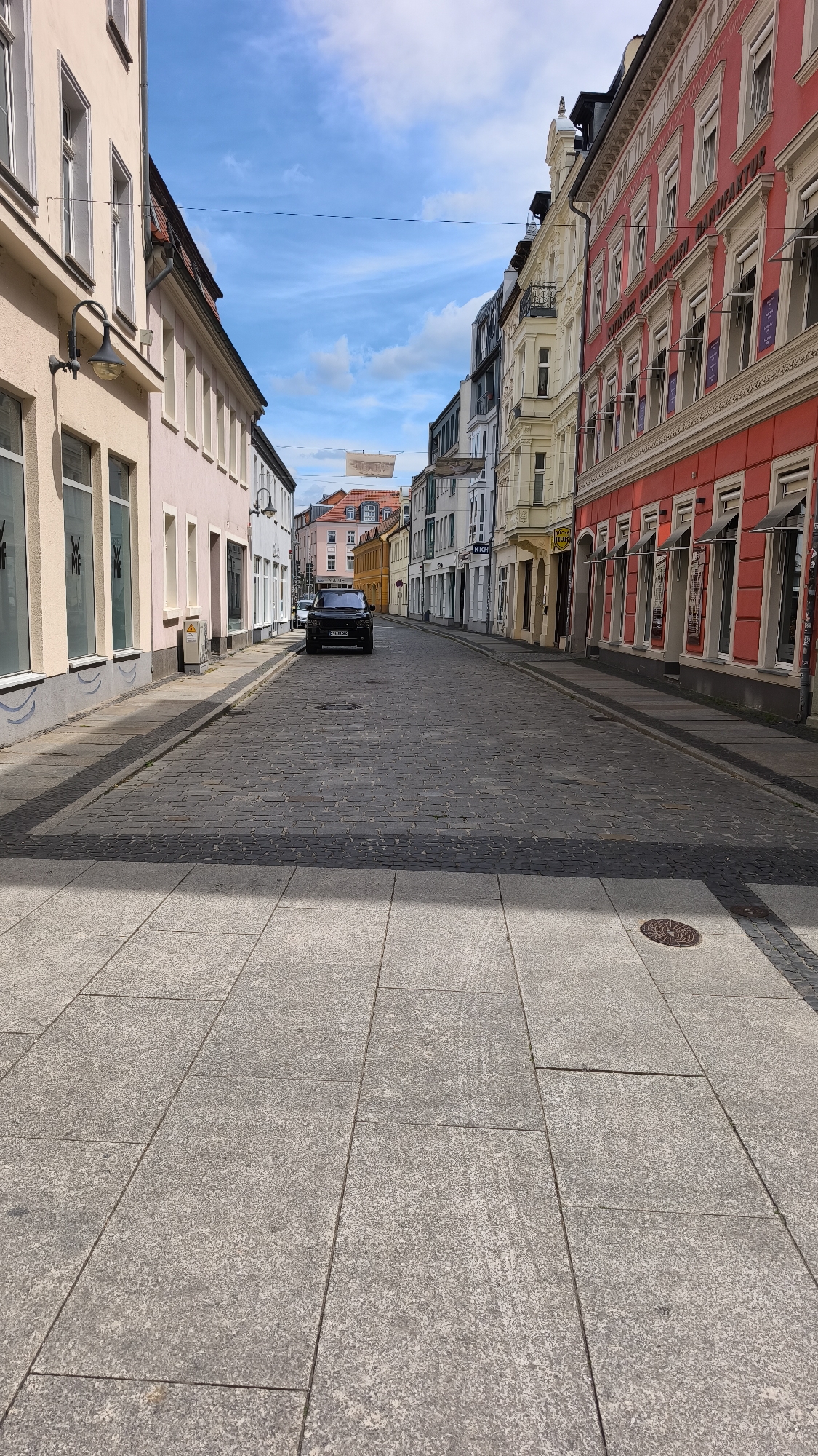 The image depicts a street scene in an unknown city. The street is paved with cobblestones. There is a black vehicle situated in the middle of the street. Buildings of varying colors and heights line both sides of the street. The sidewalk in the foreground is constructed of large, rectangular paving stones. Overhead, the sky is partially obscured by the buildings, with visible patches of blue sky and clouds.