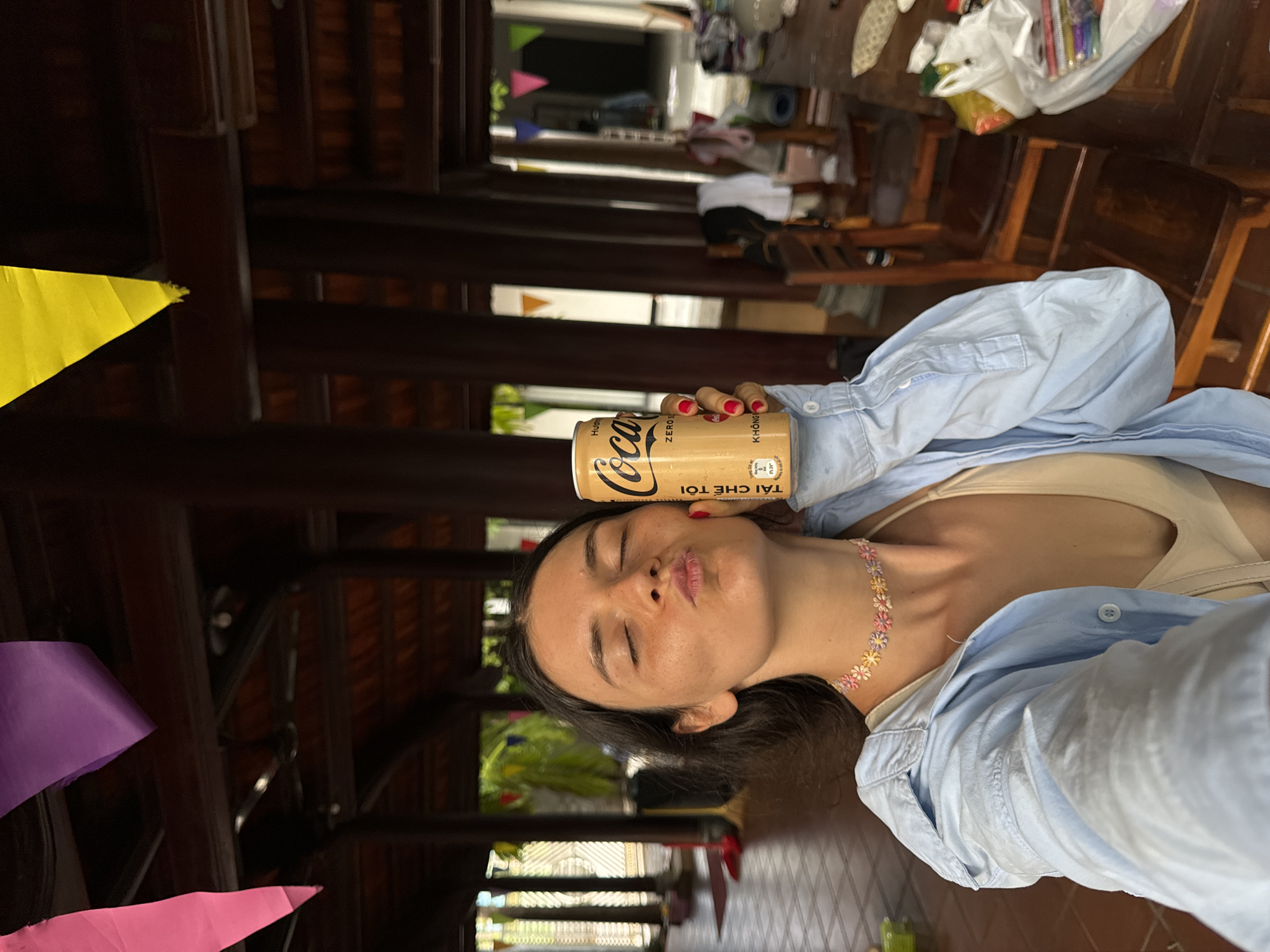 A young woman, likely in her early twenties, is the sole subject in this portrait-oriented, indoor photograph. She is captured from the chest up, with her head tilted back and her eyes closed as she puckers her lips, holding a can of Coca-Cola Zero Sugar against her forehead. She wears a light blue, unbuttoned collared shirt over a light-colored tank top and a delicate, multi-colored floral choker necklace. Her nails are painted a vibrant red.
The background suggests an indoor, possibly traditional Vietnamese setting. Dark wooden beams and architectural details are visible, hinting at a resort or a substantial building with a rustic aesthetic. To the left of the frame, colorful triangular pennants in yellow, purple, and pink are partially visible, adding a festive or decorative touch.
In the upper right portion of the image, a table is laden with various items, including plastic bags, what appears to be a yoga mat, and other miscellaneous objects. The lighting is consistent with daytime, though the specific time is not discernible. The overall mood is playful and relaxed, conveyed through the woman's gesture and expression.
The can of Coca-Cola Zero Sugar has visible text in both English and Vietnamese. The English text reads "ZERO SUGAR," and the Vietnamese text includes "HƯƠNG CHANH SẢ," which translates to "Lemongrass Flavor." Additional Vietnamese text also appears on the can. The location context of Mũi Né, Vietnam, aligns with the presence of Vietnamese text on the product and the style of the background architecture.