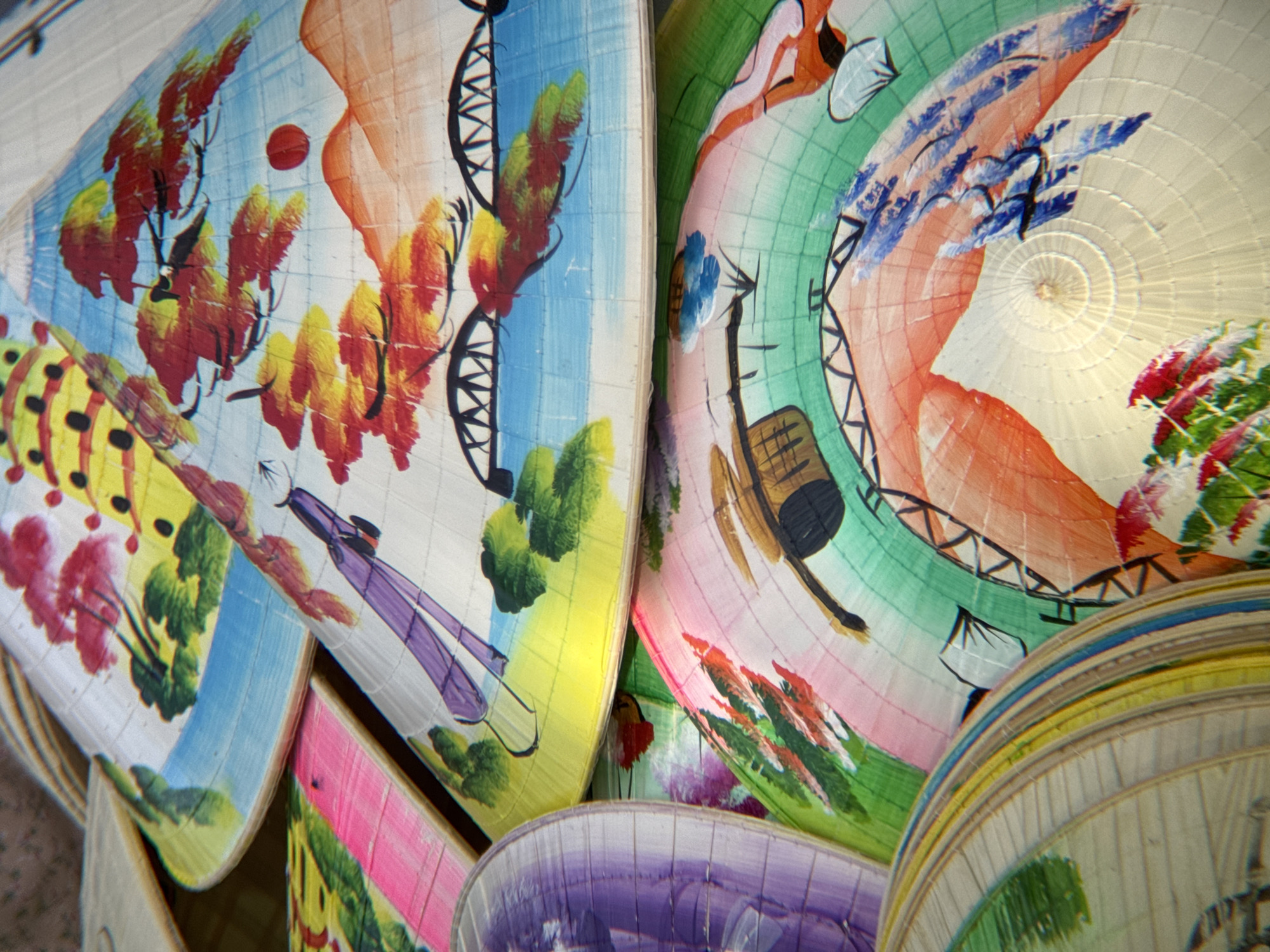 In Phú Xuân Ward, Vietnam, a close-up shot showcases a collection of vibrantly painted conical hats, known as Nón lá. The hats are stacked and overlapping, creating a layered visual effect. Each hat features a unique, hand-painted scene. One hat displays a landscape with red and yellow trees against a blue sky, with stylized mountains and a red sun. Another hat features a purple figure and green foliage. A third hat showcases a rainbow-like gradient of colors, from pink to green, with painted elements suggesting a still life or cultural motif. The intricate details of the paintings and the woven texture of the hats are clearly visible. The lighting suggests an outdoor setting, possibly a market or artisan's stall, with natural light illuminating the colorful designs.