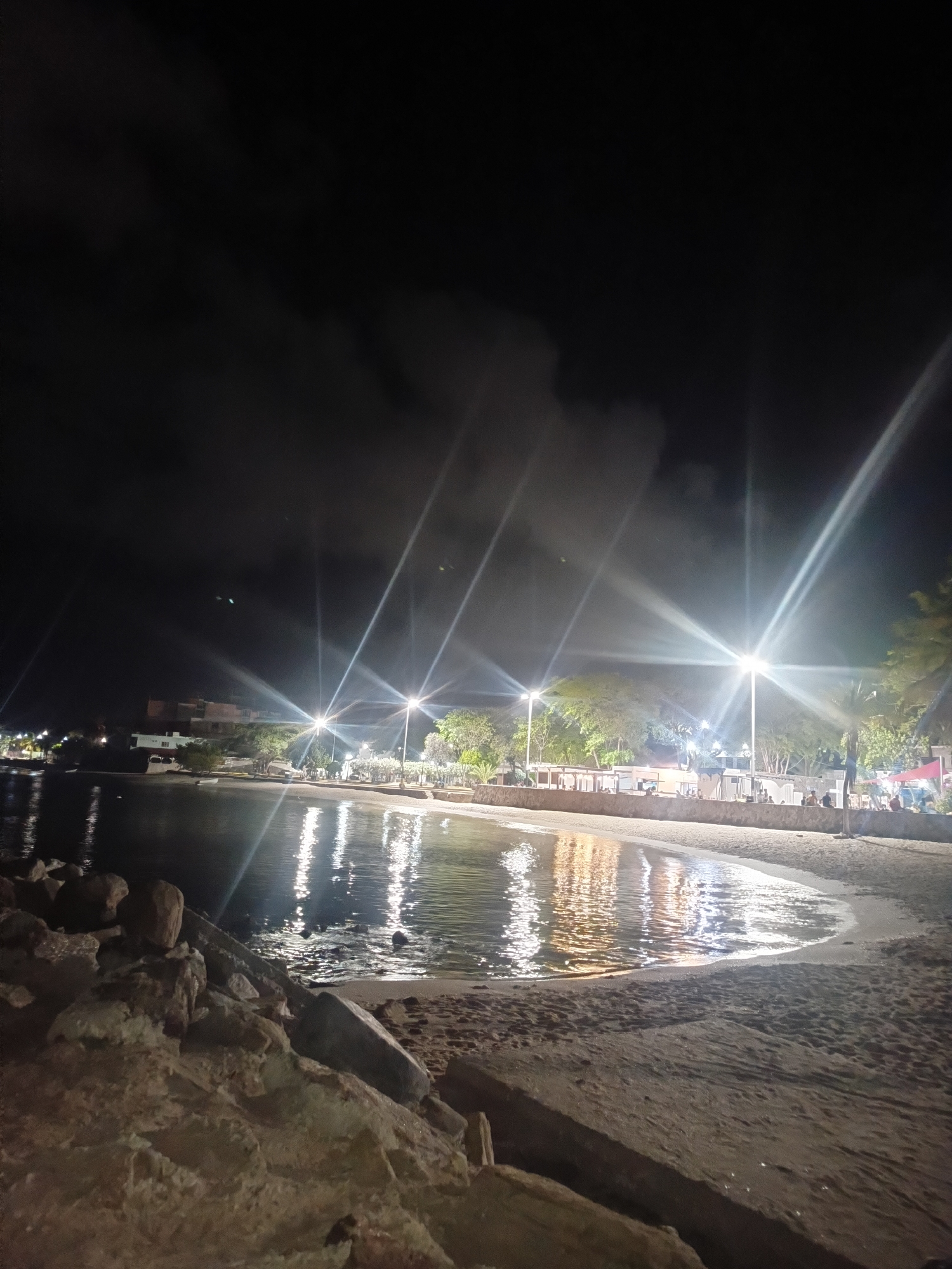 The image depicts a nighttime scene on a beach in Juan Griego, Venezuela. In the foreground, large, weathered rocks line the edge of the sandy beach. The water's edge is visible, with gentle waves lapping onto the shore. Beyond the beach, a promenade or walkway is illuminated by streetlights, casting long, bright streaks across the scene. Buildings, possibly restaurants or shops, are visible behind the promenade, with lights illuminating their facades. Several trees are scattered along the promenade area, adding to the ambiance. A few people can be seen walking or sitting along the promenade, enjoying the evening. The sky is dark, with some faint clouds, and the overall lighting suggests it is nighttime. The reflection of the lights on the water creates a shimmering effect.