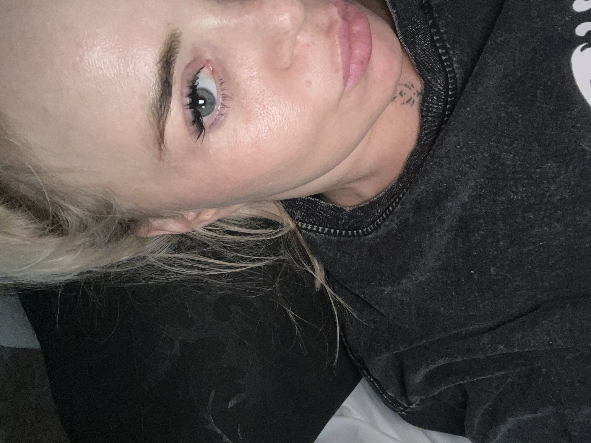 A close-up, overhead shot shows a young woman with blonde hair and light skin lying down. She is wearing a dark grey t-shirt. Her hair is pulled back loosely, with some strands falling forward. Her right eye is visible, showing blue iris and dark eyelashes. Her mouth is slightly open, revealing pink lips. A small, dark tattoo is visible on her neck, below her jawline. The background is dark and out of focus, with a hint of white fabric on the left. The lighting is dim, creating soft shadows on her face.