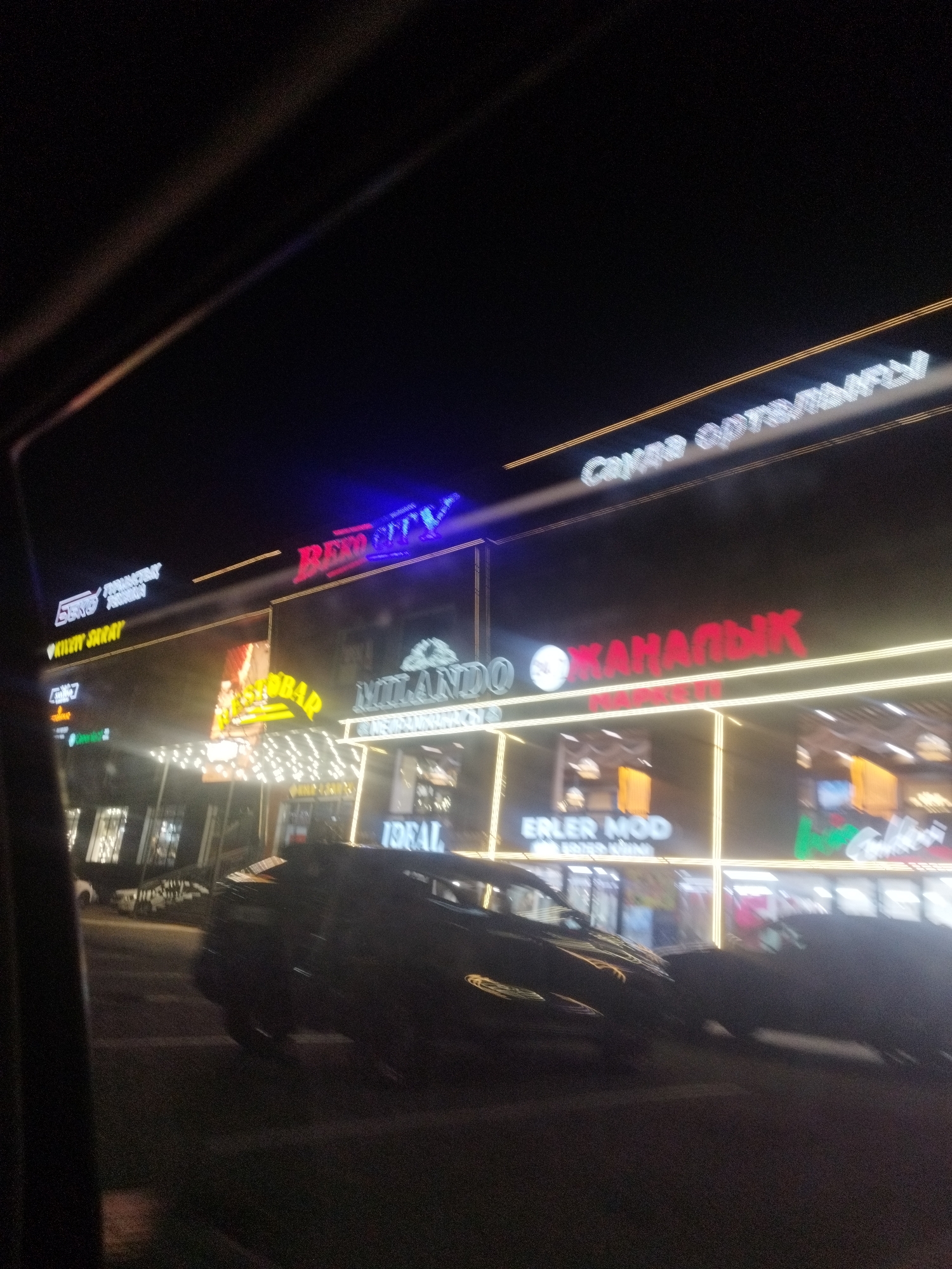 The image shows a street scene at night in Shymkent, Kazakhstan, likely in a commercial district. A dark-colored car is parked or moving on the street in the foreground. In the background, several brightly lit signs of shops and businesses are visible. Some of the signs are in Cyrillic script, such as "RED CITY," "MILANDO," and what appears to be "ЖАНАЛЫК." Another sign reads "ERLER MOD." The buildings are illuminated by neon lights and architectural lighting, creating a vibrant but blurry effect due to the low light conditions and motion blur. The overall impression is that of a lively nighttime shopping or entertainment area.