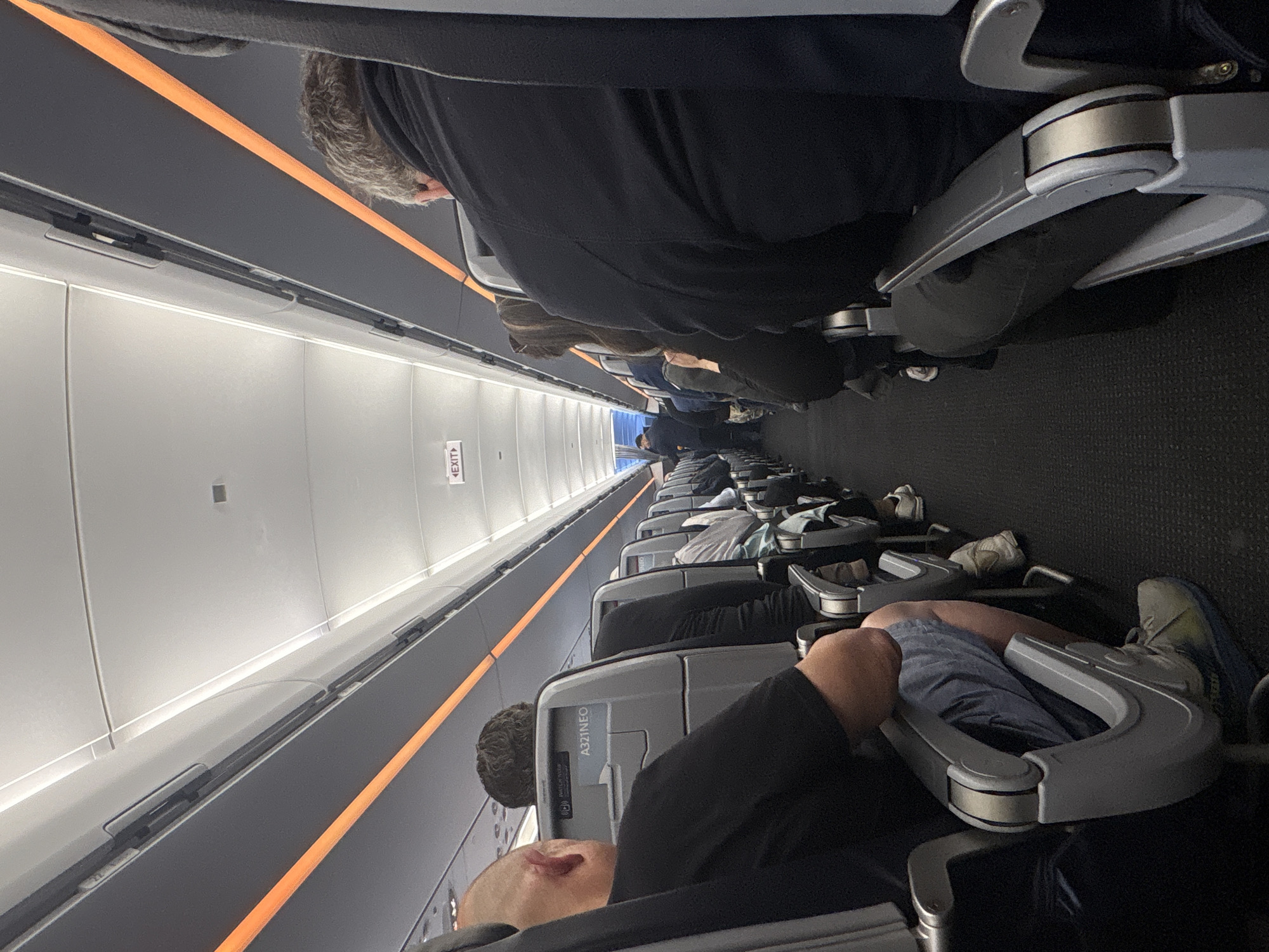 The image shows an airplane cabin interior. Several rows of seats are visible, occupied by passengers. Overhead compartments line the cabin above the seats. The plane is traveling to or from New York.