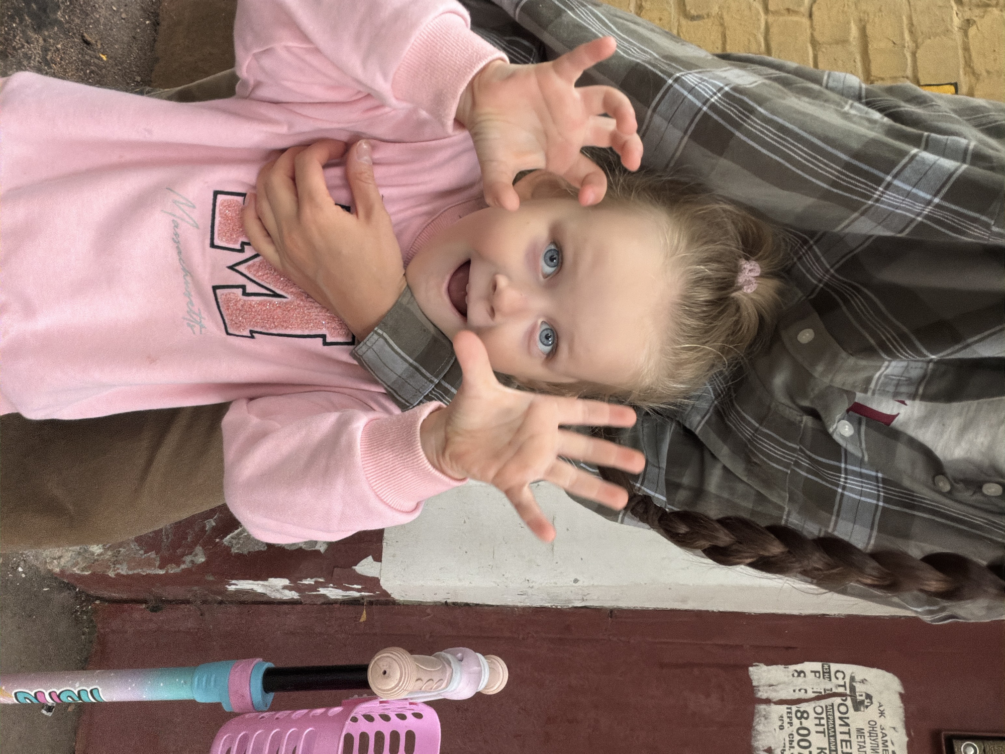 A close-up shot captures a young girl with bright blue eyes and blonde hair pulled into a ponytail, wearing a pink sweatshirt with the letter "M" and "Massachusetts" printed on it. She is looking at the camera with an open mouth and wide eyes, raising her hands in a playful gesture. An adult's arm is visible around her shoulders, and their hand rests on her chest. Behind the girl, a person's dark brown braided hair hangs down their back, and they are wearing a grey and white plaid shirt. To the right of the girl, part of a pink and blue scooter with a pink basket is visible. The background features a textured yellow brick wall on the left, and a reddish-brown wall with peeling paint and some torn advertisements on the right. The ground appears to be asphalt or concrete. The scene is set in Malakhovka, Russia.