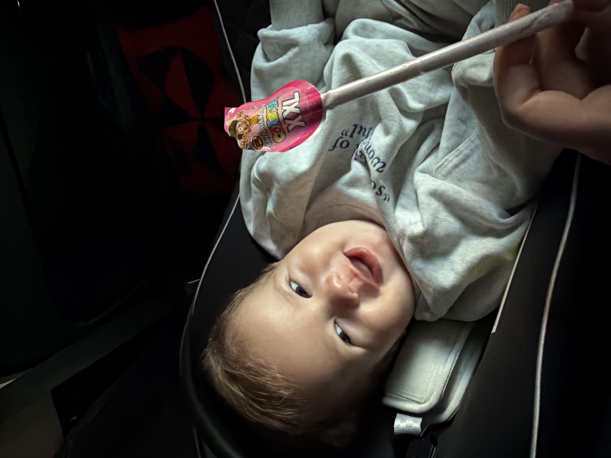 A baby is lying in a car seat, held by a person's hand, and is offered a lollipop. The baby has blond hair and is wearing a gray hoodie. The lollipop has a pink wrapper with a cartoon character on it and the text "XXL". The background is dark, with a red and black pattern visible in the upper right corner. The scene appears to be inside a car. This image is not associated with the city of Kazan, Russia.