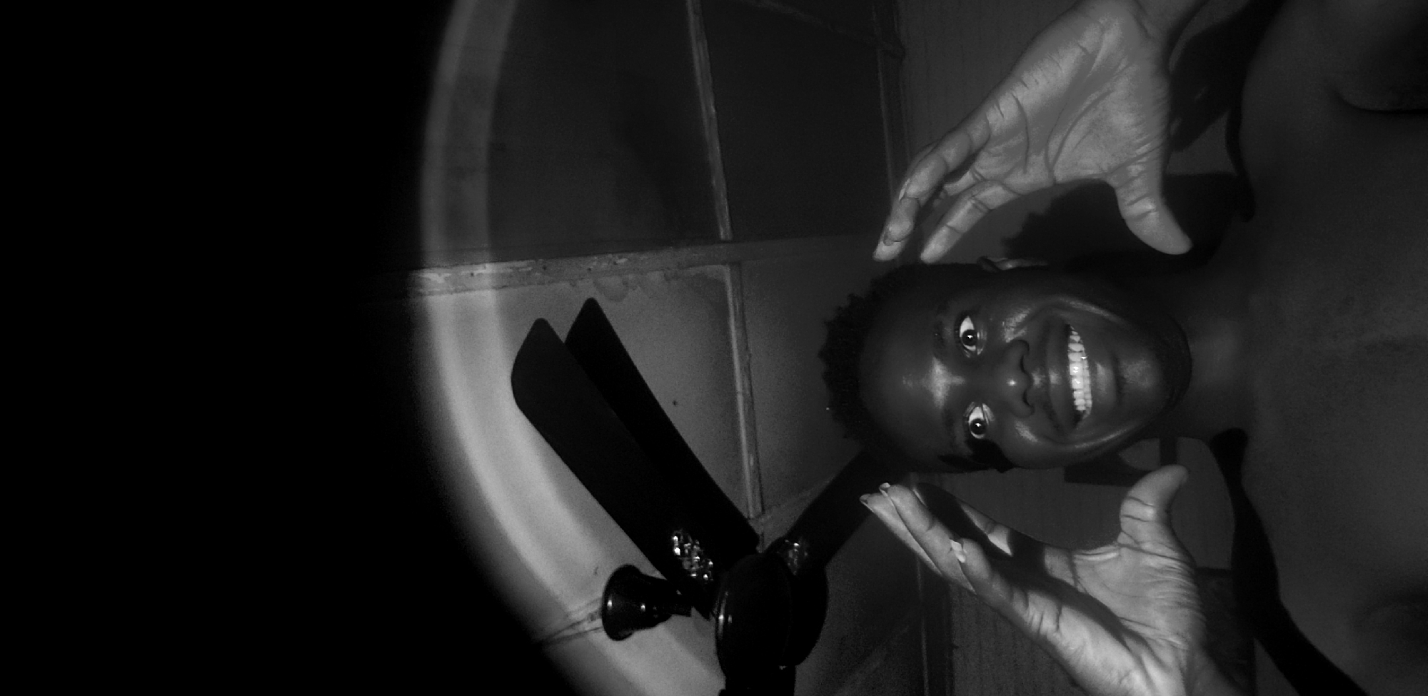 In a dimly lit interior, a Black male subject, shirtless, is pictured from the chest up. He is looking directly at the camera with wide, startled eyes and a broad smile, his teeth prominently displayed. His hands are raised to either side of his head, fingers splayed, as if in a gesture of surprise or exasperation. Above and behind him, a ceiling fan is visible with its blades angled. The lighting creates stark contrasts, casting shadows and highlighting the textures of his skin and the objects in the environment. The background appears to be a ceiling with visible structural elements. The location of Narayi, Nigeria, is noted for this scene.
