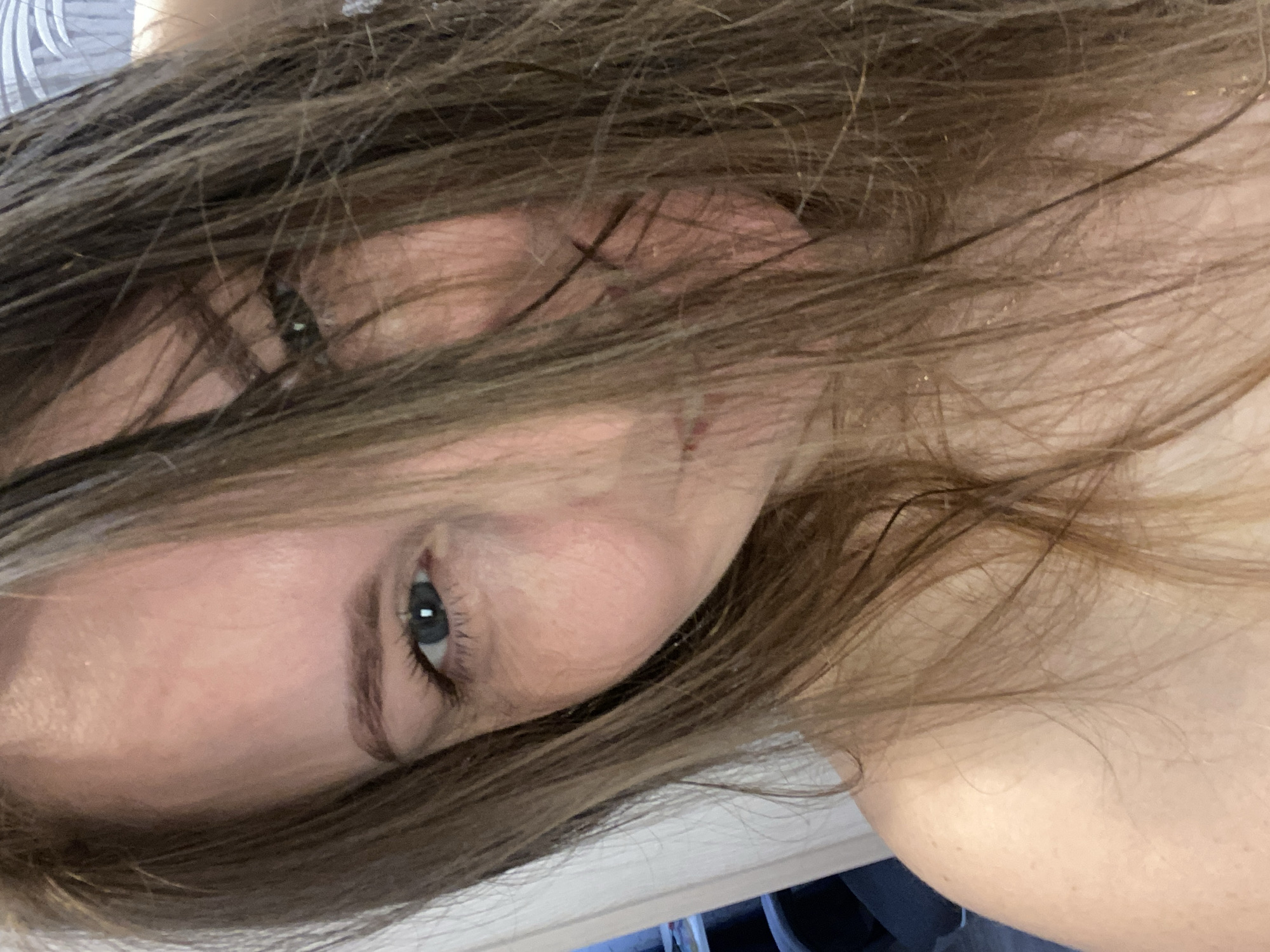 A close-up portrait of a woman's face, tilted diagonally. She has fair skin, brown hair partially obscuring her face, and a visible blue eye. The background shows a glimpse of a white surface and dark objects, possibly furniture. There is no indication of a specific location or reference to Noviy, Russia.