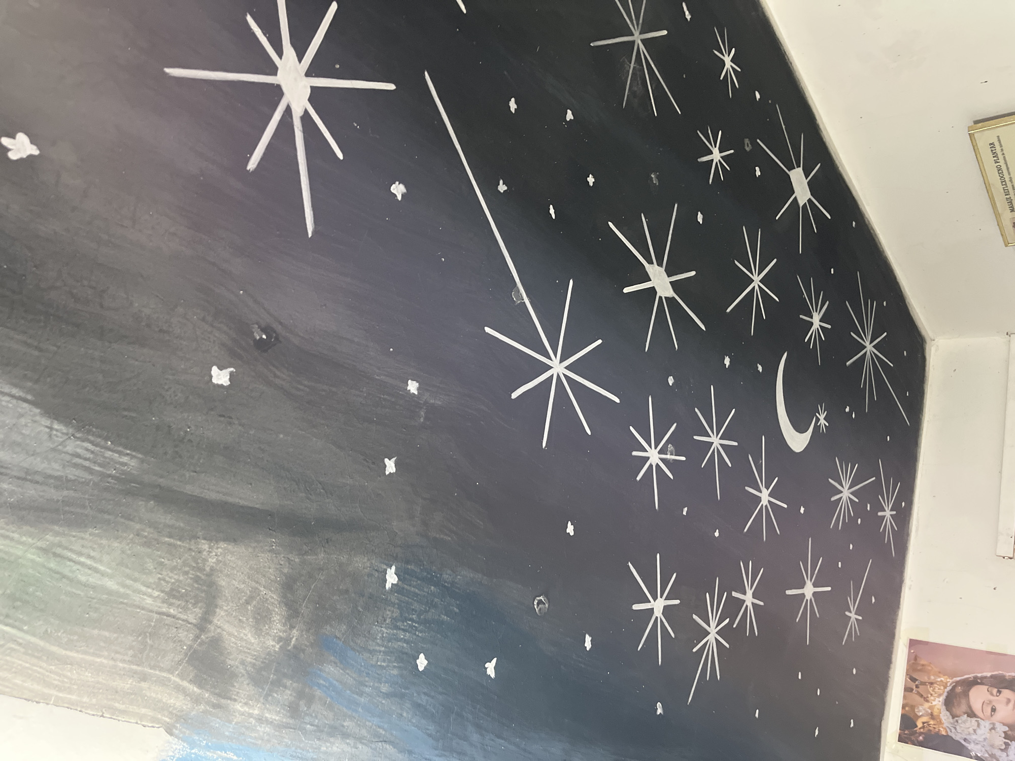 The image shows a wall painting in Juan Griego, Venezuela. The painting depicts a night sky scene with a dark, gradient background representing the sky. Numerous white stars of varying sizes and shapes are painted on the wall, along with a crescent moon. A shooting star is also depicted with a long, white line extending from a star. The painting appears to be on an interior wall, possibly in a residential or public space. A framed document and a photograph are partially visible on the wall to the right of the painting. The wall appears to be in a corner where two walls meet the ceiling. The painting shows signs of wear and tear with visible paint strokes and surface imperfections.