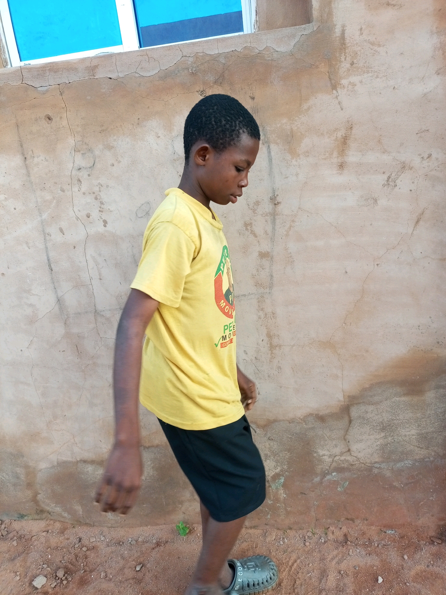 In Tempure, Nigeria, a young boy in a yellow t-shirt and black shorts walks along a cracked, beige concrete wall. Above the wall, blue-tinted windows are visible. The ground is a mix of sand and small pebbles, with a few green plants growing near the wall. The boy is in profile, looking down as he walks. The t-shirt has a circular logo with text that includes "PEACE" and "MOVE."