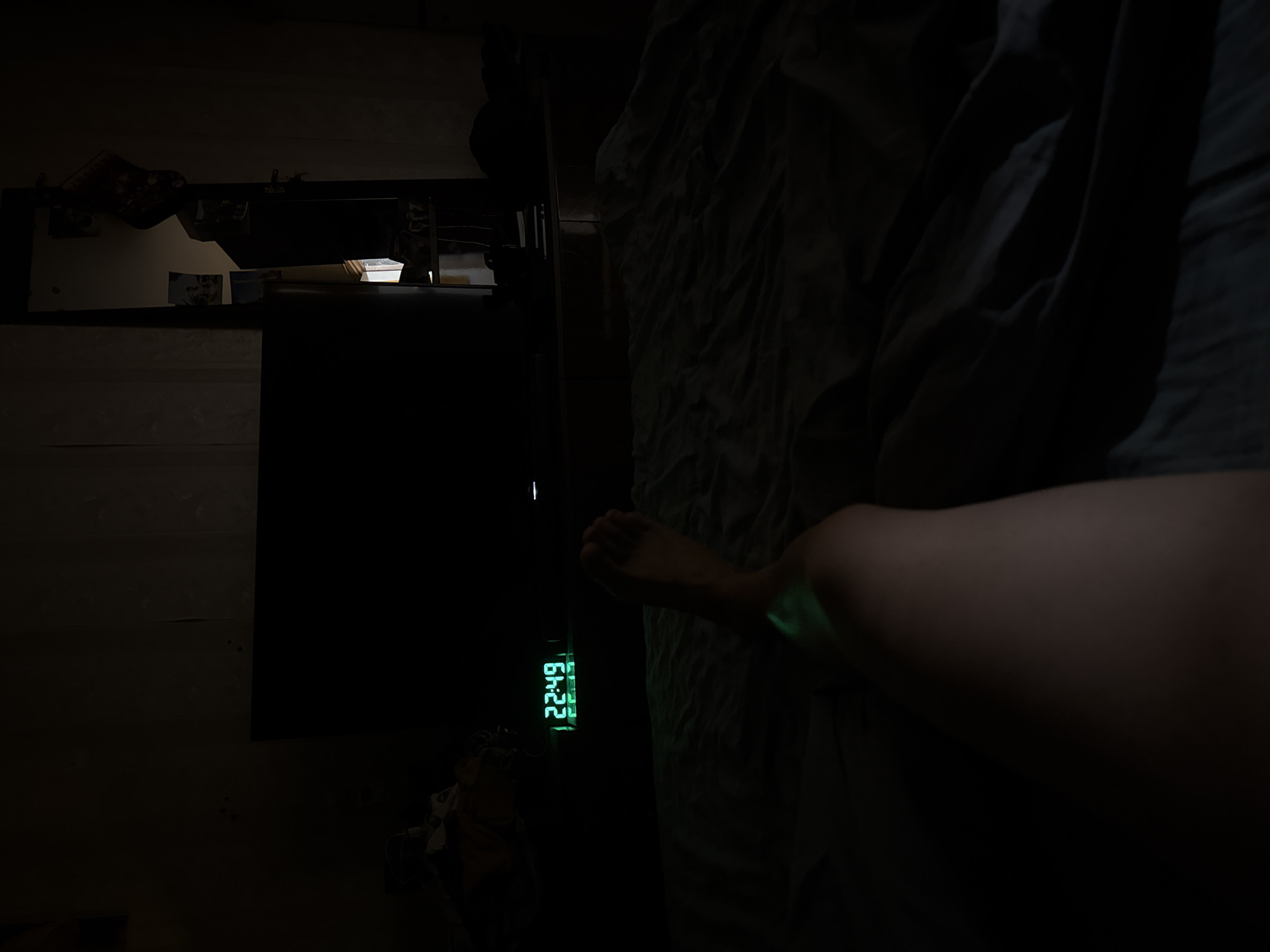 The image shows a dimly lit room, presumably in a residential setting. The visible elements include a bedside table with various items on top, a digital clock displaying the time 22:49 with a green glow, and a person lying in bed. The background appears to be a wall. There is not enough information to confirm the location of the room in Khimki, Russia.