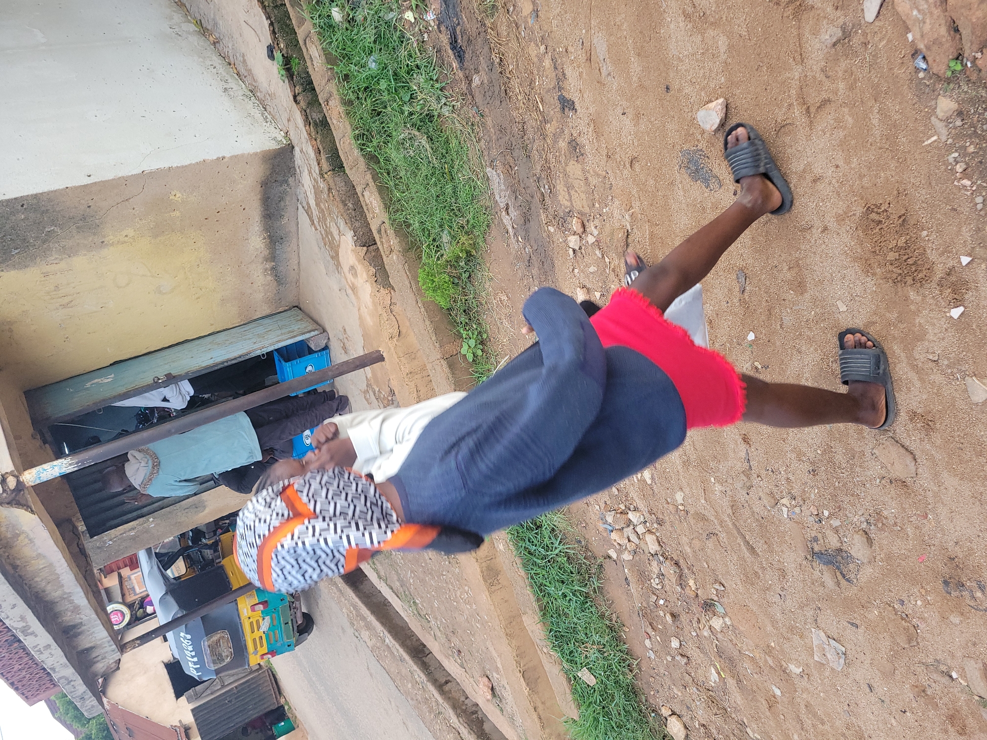 The image presents an outdoor street scene in Jos, Nigeria, featuring a combination of structures, individuals, and an unpaved ground surface. In the mid-foreground, a person with dark skin is depicted in motion, walking rightward. They wear a dark blue long-sleeved top, red shorts, and black slide sandals. A head covering with a white, black, and orange geometric pattern is visible on their head. A partial view of another person's arm, clad in a white sleeve, is adjacent to the foreground figure.
In the background, a two-story building with a light yellow, weathered, and cracked facade features an open, light blue metal door. Within or near this entrance, two individuals are observed. One person, viewed from behind, wears a light teal hooded top with a textured or fur-lined trim on the hood, paired with dark trousers. The second individual is partially visible, wearing a white top. Several blue plastic crates, one marked "NB," are positioned near the building's doorway.
On the paved road behind the foreground, a black and yellow three-wheeled auto-rickshaw (Keke Napep) with teal accents is parked. The vehicle displays the alphanumeric sequence "PF122JJ" on its rear panel. To the left and further back, another building with a reddish-brown roof and a dark entrance is visible.
The ground in the foreground and mid-ground is composed of reddish-brown sandy soil, interspersed with scattered rocks, pebbles, and darker, moist patches of earth. A concrete drainage channel or gutter runs along the left side, accompanied by patches of green grass and minor debris. A dark, slender metal pole extends vertically from the lower left of the frame. The scene is illuminated by natural daylight.