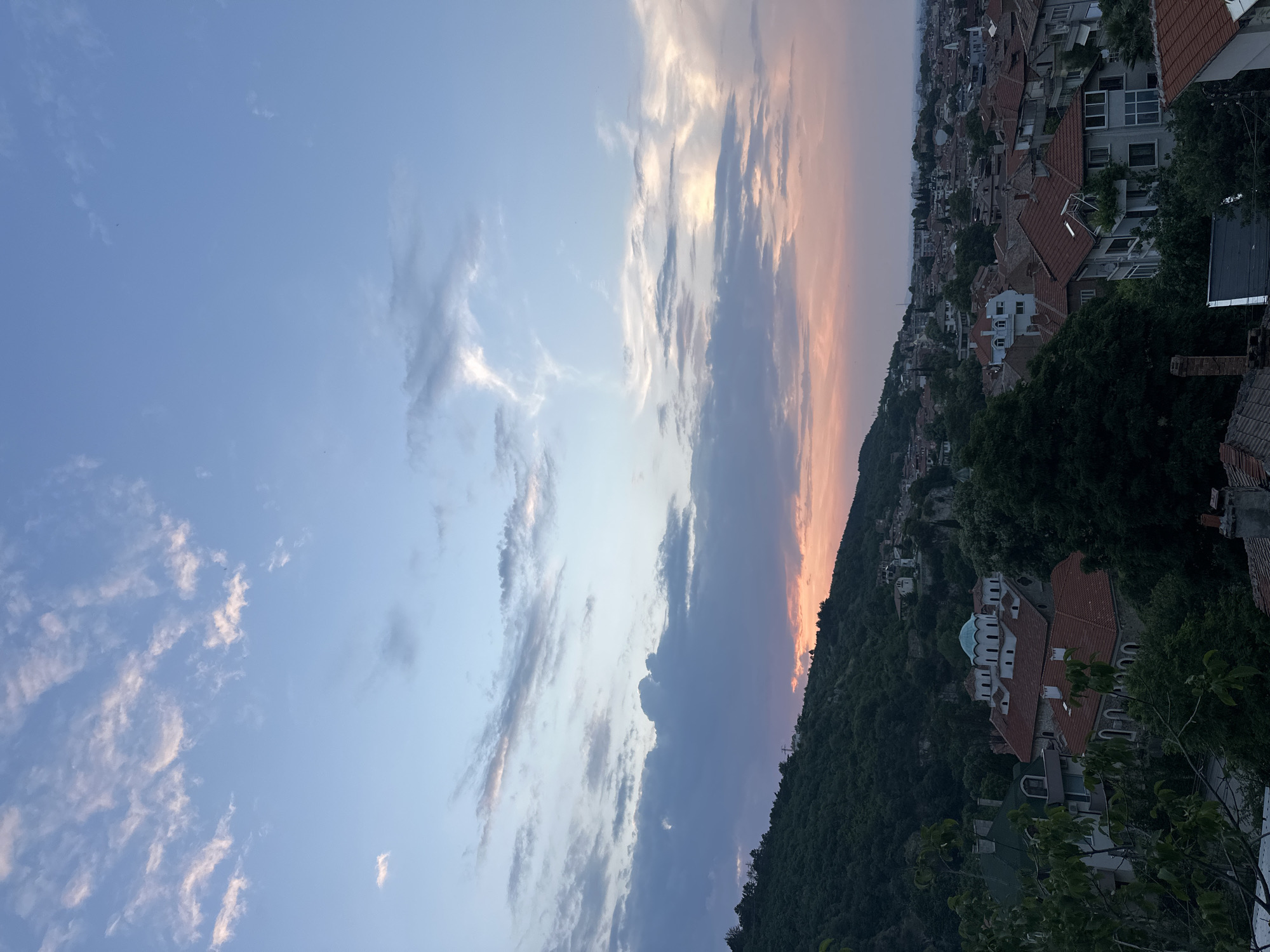 The image presents a detailed panoramic view of the city of Asenovgrad, Bulgaria, captured during twilight or sunset from an elevated vantage point. The upper half of the frame is dominated by a expansive sky, transitioning from a light blue at the zenith to soft pink and orange hues along the horizon on the right, indicative of the setting sun. Various cloud formations are visible: scattered, wispy white clouds are present in the upper left, while denser, darker grey clouds accumulate in the mid-left, gradually becoming more illuminated with warm light towards the right side of the horizon. A few minute, dark specks, consistent with distant birds, are discernible against the clear blue sky in the upper left quadrant.
The lower half of the image features a prominent, heavily forested hill on the left, densely covered with dark green trees, its upper ridge line dimly silhouetted against a subtle reddish-orange glow from the sunset. This natural landscape merges into the urban environment of Asenovgrad. The city consists of a dense arrangement of residential buildings, predominantly characterized by their numerous reddish-brown terracotta-tiled roofs. Situated towards the center-left of the urban area, nestled amidst trees, is a distinctive large building identified as a church or monastery. It features multiple domes, several of which exhibit a light teal or turquoise coloration, complementing its traditional tiled roofs. The urban sprawl extends across the lower and right portions of the image, receding into the distant haze. No individuals are present or discernible within the scene, and no specific actions or interactions are depicted. The lighting is soft and diffused, casting gentle, indistinct shadows across the landscape and urban structures.
