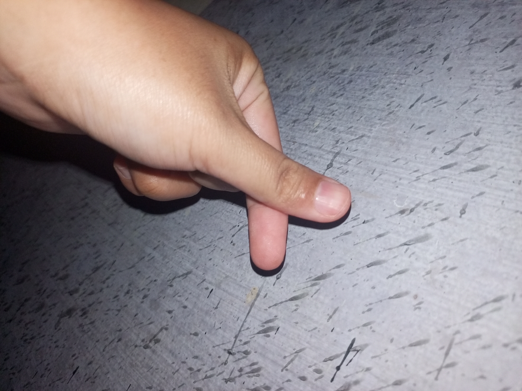 A human hand is positioned above a grey, speckled surface. The index finger is extended downwards, its tip almost touching the surface. The hand and finger appear to be the primary subjects. The background is an abstract, textured surface, possibly concrete or asphalt, with irregular dark marks scattered across it. The lighting suggests an indoor or dimly lit outdoor environment. The image does not contain any information about the city of Sei Beras Sekata, Indonesia.