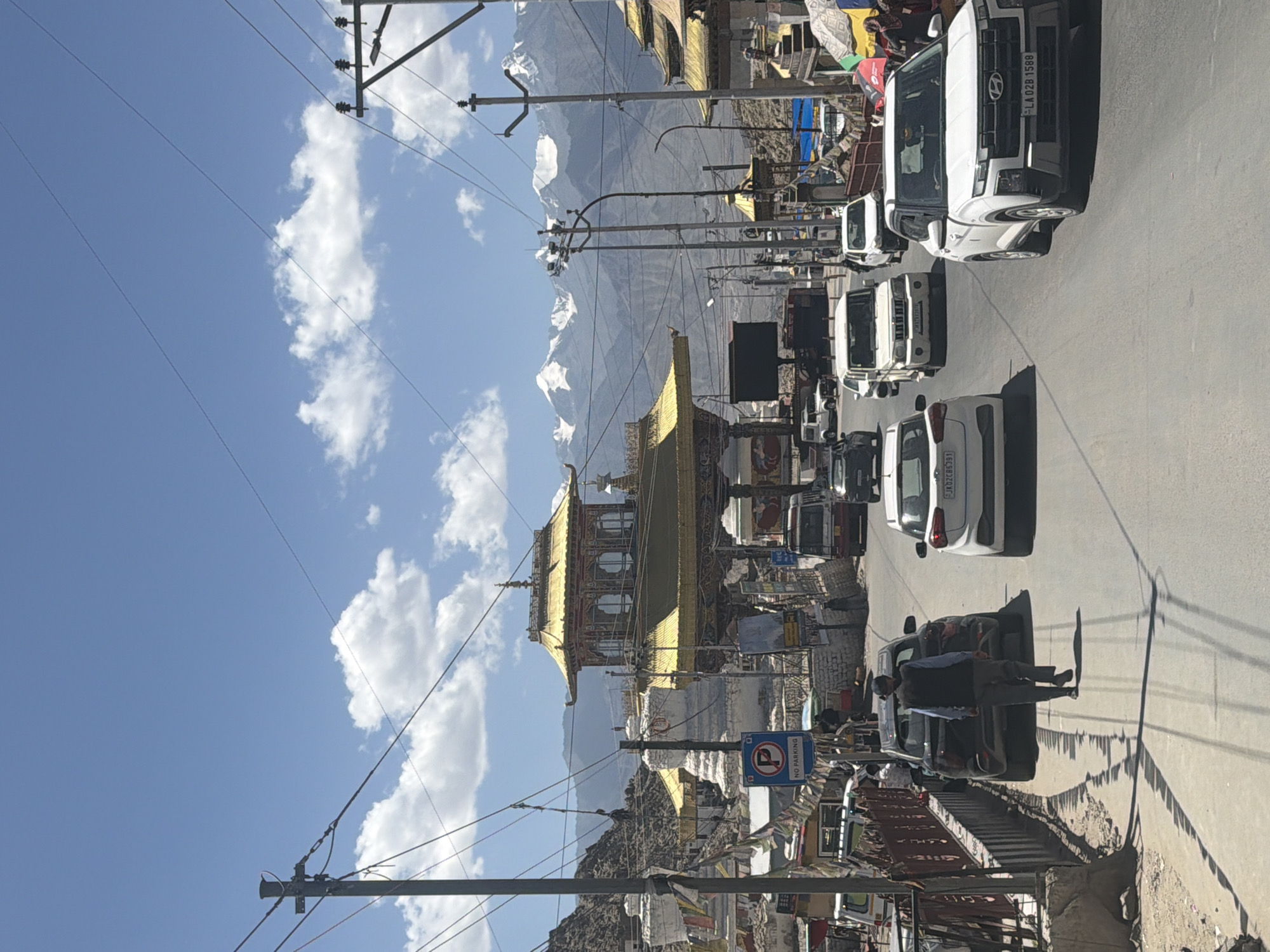 The image depicts a street scene in Leh, India, with a prominent golden-roofed structure resembling a temple or gateway in the background. Several cars are visible on the road, with a man walking away from the camera on the left side of the street. Snow-capped mountains are visible in the distance under a blue sky with scattered clouds. The architecture suggests a Tibetan or Buddhist influence, consistent with Leh's location in Ladakh. There are several utility poles with overhead wires, indicating an urban or semi-urban setting. The overall atmosphere is bright and sunny, suggesting a daytime scene.