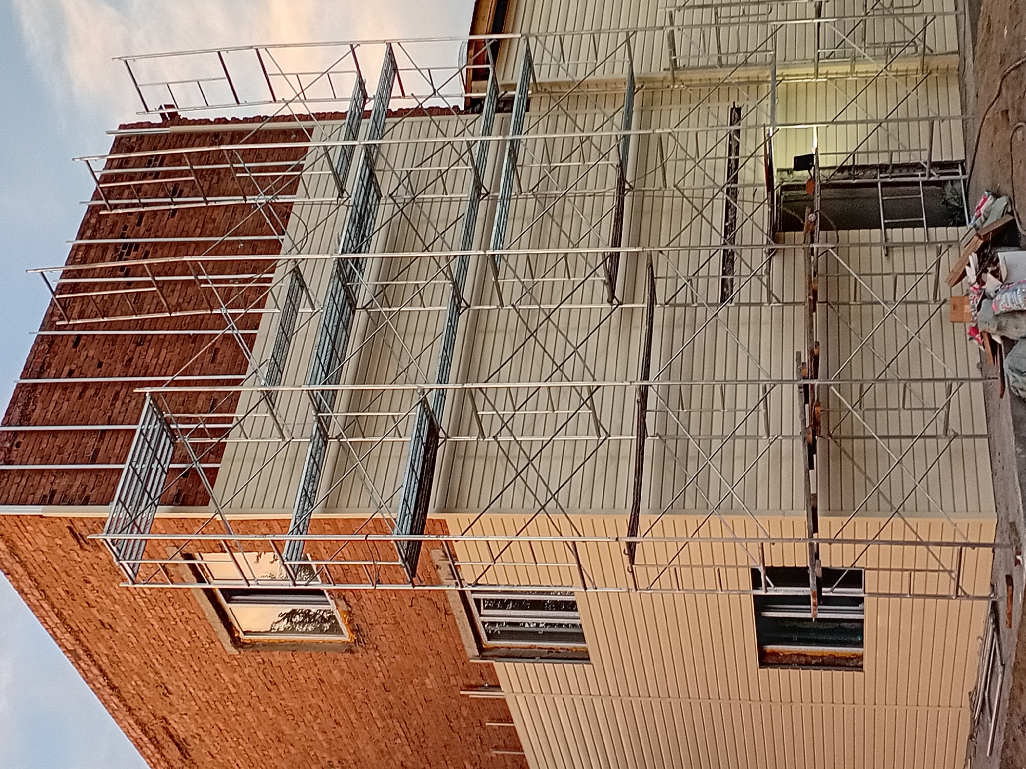 The image depicts a building under construction in the city of Semey, Kazakhstan. The structure features a brick upper section and a beige siding lower section. Extensive scaffolding is erected around the building, suggesting ongoing renovation or new construction work. Several windows are visible on both the brick and siding portions of the building. The ground in front of the building is covered with dirt and construction debris, including wood planks and bags. The sky in the background is overcast with clouds. No people are visible in the image.