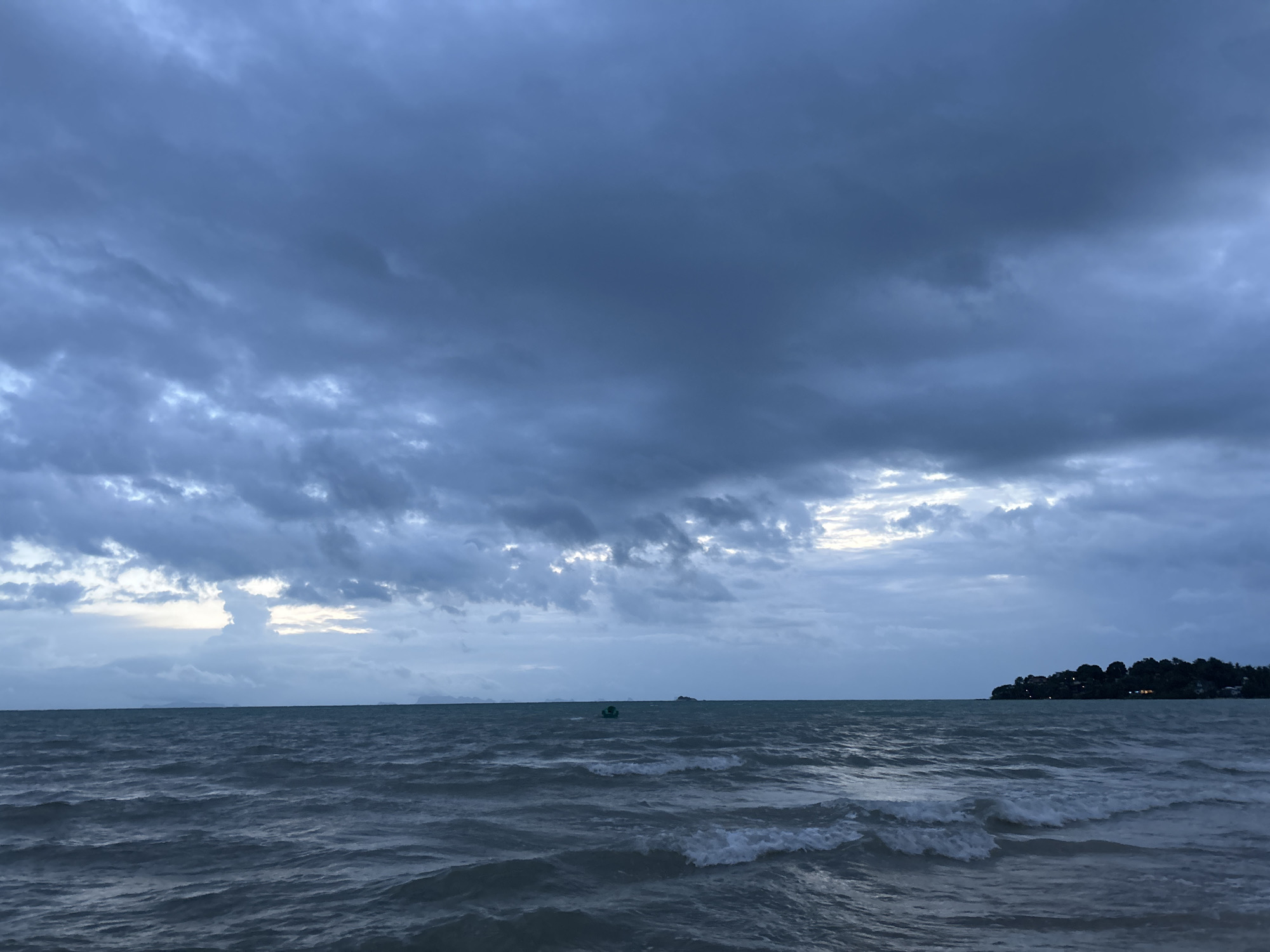 A wide shot shows a turbulent ocean under a cloudy, overcast sky. The sky is filled with dark grey clouds, with some lighter patches of sky visible near the horizon. The ocean is a deep blue-grey, with white-capped waves rolling towards the foreground. A small, dark green boat is visible in the distance, as is a tree-covered island with a few buildings on the right side of the frame. The scene appears to be a coastal area in Ko Pha-ngan District, Thailand, likely during a period of inclement weather. There are no people visible in the image.