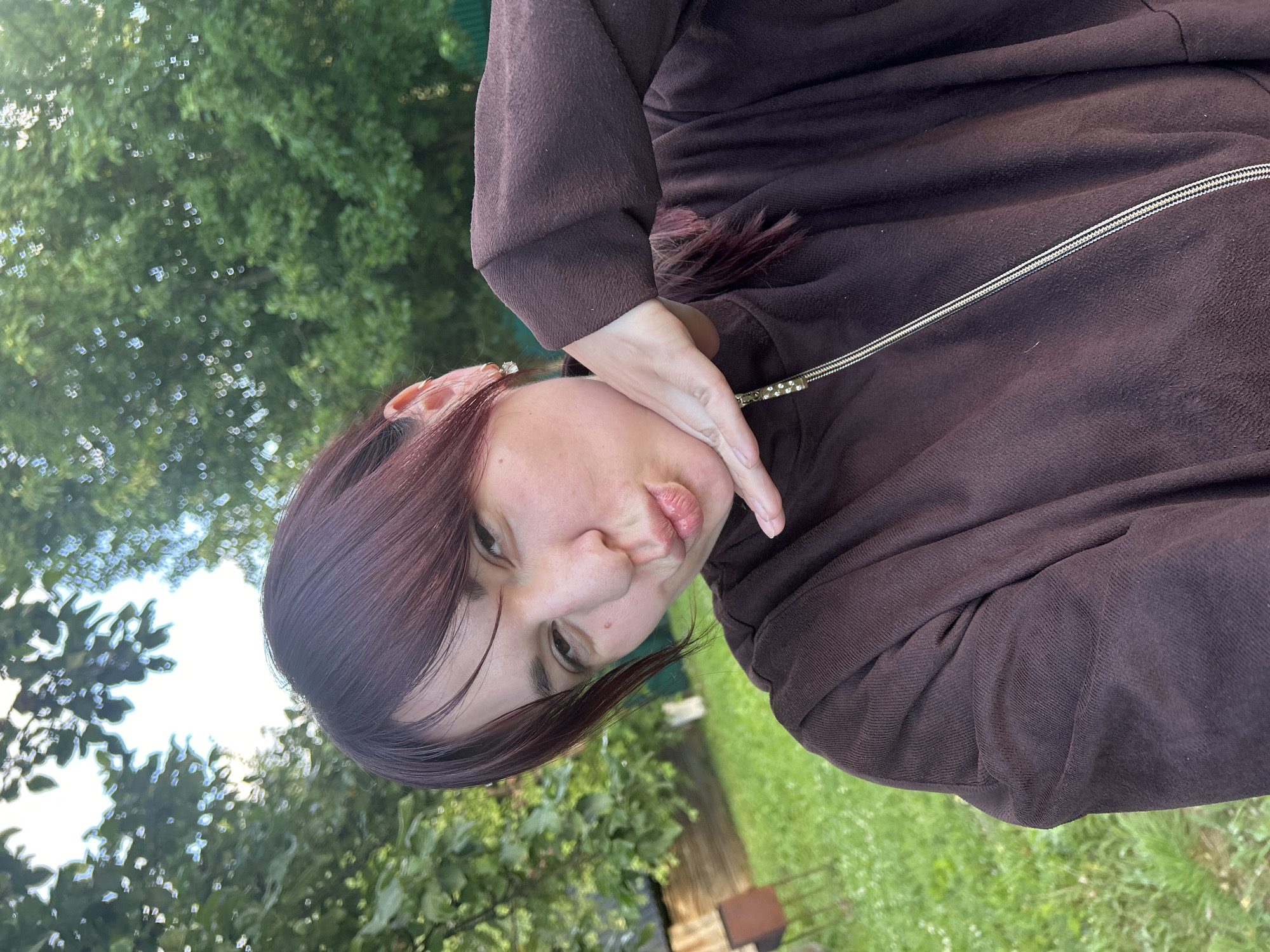 The image shows a young woman with dark red hair and bangs, pouting her lips and holding her hand near her chin. She is wearing a dark brown zippered hoodie. She has a small, square earring in her visible ear. The background is an outdoor setting with green grass, trees, and a wooden structure that could be a shed or a small house. The sky is visible through the leaves of the trees. The location is identified as Фоки, Russia.