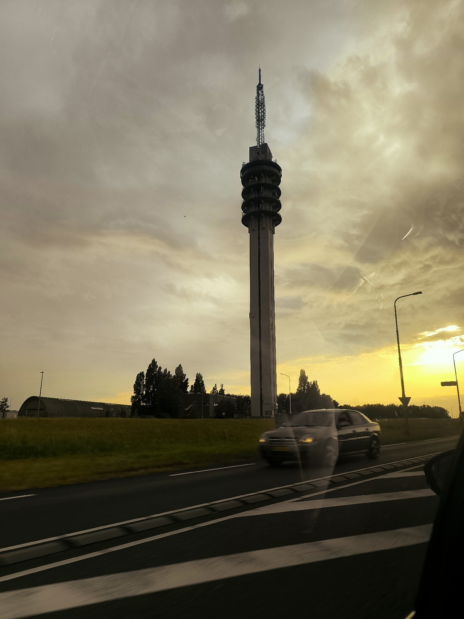 The image depicts a view from a moving vehicle in Lelystad, Netherlands, with the iconic Telecommunication Tower of Lelystad (also known as the KPN Tower) as the central subject. The tower, a tall, concrete structure with an antenna at its apex, dominates the skyline against a dramatic, cloudy sky tinged with the warm glow of sunset or sunrise. In the foreground, the road is visible with white lane markings, and a dark-colored car is captured in motion, slightly blurred due to the vehicle's movement. The roadside features a grassy embankment, trees, and buildings, including a large, curved structure resembling a hangar. Streetlights are also present along the road. The overall impression is one of a vast, open landscape under a striking sky, characteristic of the Flevopolder region.