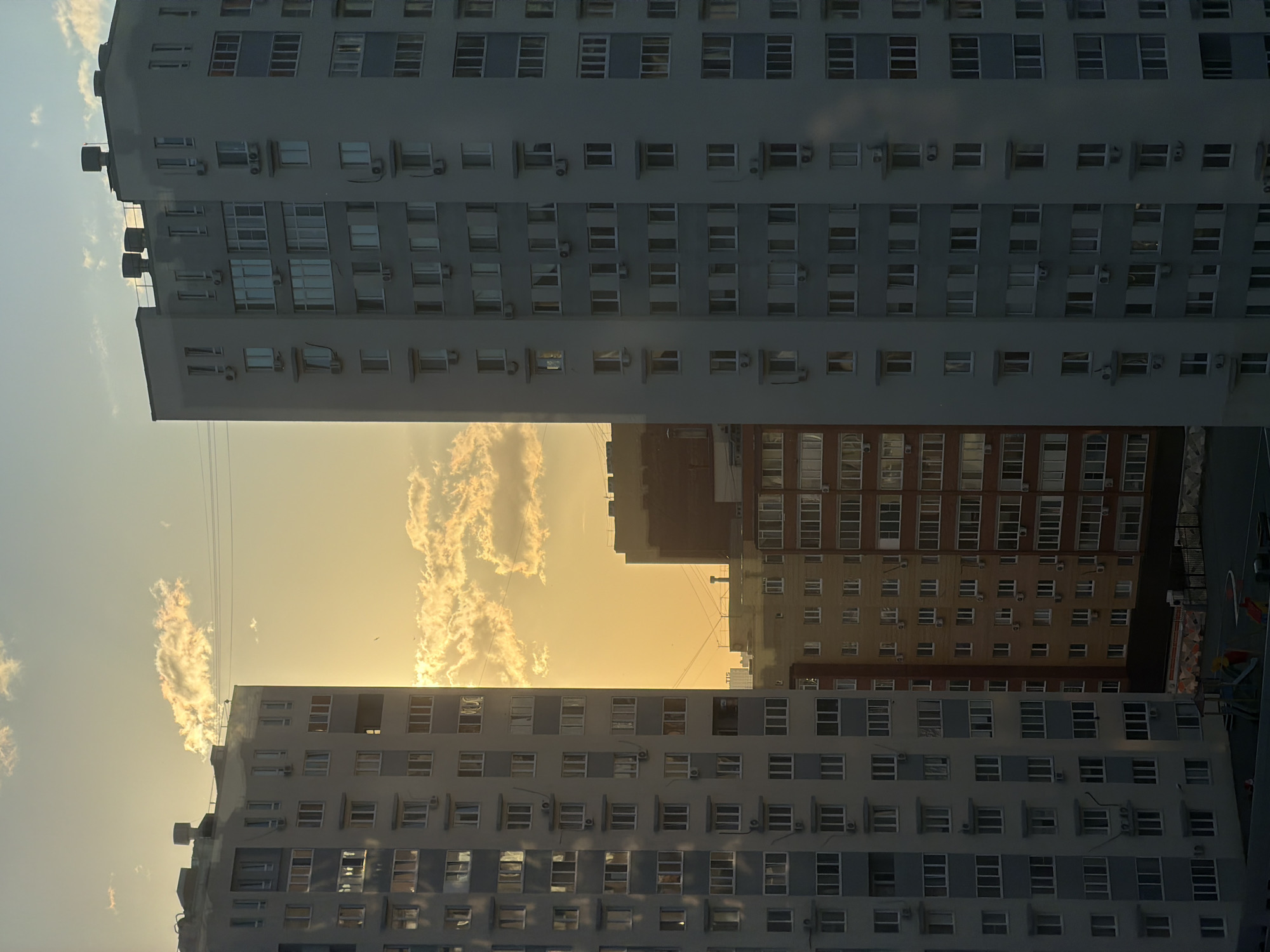 The image shows an upward view between buildings in Tyumen, Russia, likely taken during sunset. Tall, multi-story buildings with repetitive window patterns frame a golden sky with wispy clouds. The buildings are light grey and off-white. Some have external air conditioning units. Other buildings, predominantly brown/red brick are visible further away. The image is vertically oriented.