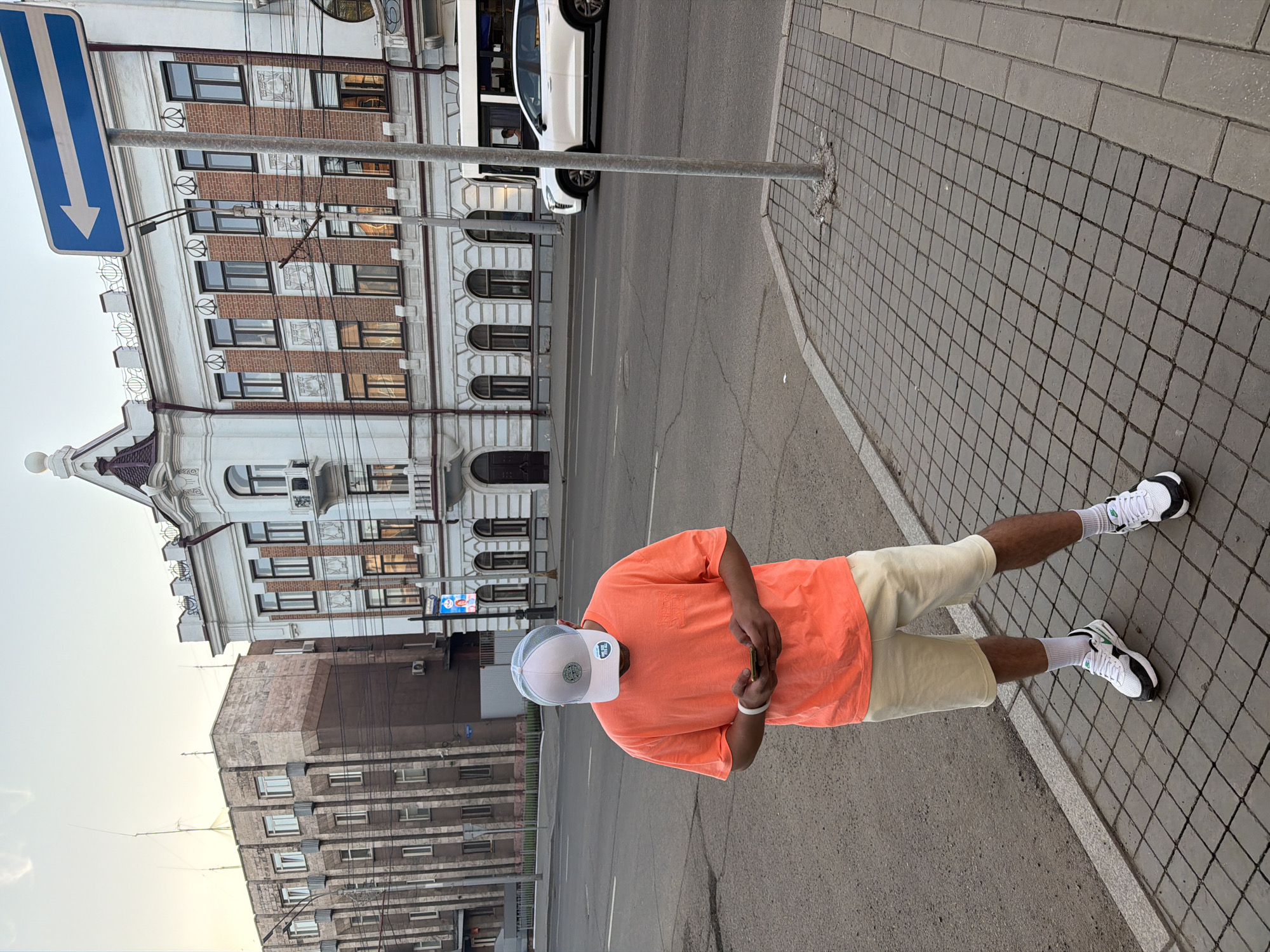 The image captures a street scene in Krasnoyarsk, Russia, under what appears to be a clear daytime sky. The primary subject is a person standing on a tiled sidewalk, holding and looking down at a smartphone.
The individual is wearing a short-sleeved, bright orange t-shirt, light beige shorts, white ankle socks, and white and black athletic shoes. They also have a white and grey baseball cap on their head and a white wristband on their left wrist. Their face is obscured as they are focused on their phone.
In the background, to the left, stands a stately multi-story building with an ornate white facade, interspersed with sections of red brickwork and featuring arched windows on its lower floors. Decorative elements adorn the roofline and window frames. Further back and to the right, a less ornate, grey and brown multi-story building with multiple windows is visible. A white SUV is partially seen on the asphalt road that runs between the sidewalk and the buildings.
A tall metal pole is positioned on the sidewalk near the person. A blue rectangular street sign with a bold white arrow pointing downwards is also visible, typically indicating traffic direction. On a small blue sign attached to the ornate building in the background, the Russian word "МЕБЕЛЬ" (MEBEL), meaning furniture, is partially legible. The sidewalk is paved with dark grey square tiles and lighter grey rectangular bricks along the curb.