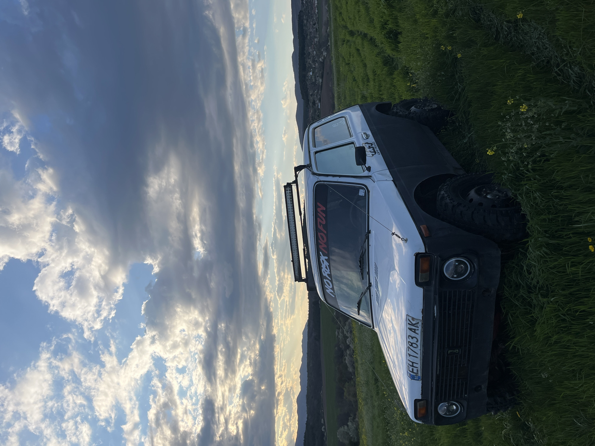 A white Lada Niva off-road vehicle, bearing Bulgarian license plate "EH 1783 AK", is parked on a grassy hillside in Varbeshnitsa, Bulgaria. The vehicle has modifications including a roof-mounted light bar and off-road tires. A sticker reading "NO REX NO FUN" is visible on the windshield. The background consists of a cloudy sky, a distant hillside with a scattering of buildings suggesting a village or urban area, and grassy terrain.