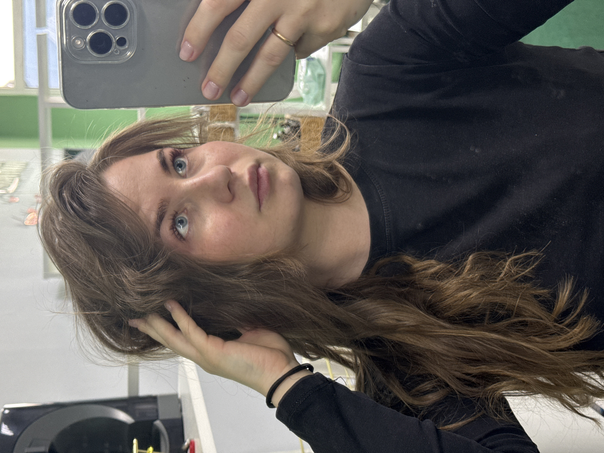 A young woman with fair skin, long brown hair, and blue eyes is taking a selfie. She is wearing a black long-sleeved shirt. Her hair is styled with loose curls, and she is touching it with her left hand. She is holding a silver smartphone with a multi-lens camera in her right hand, which has a gold ring on the ring finger. The background is slightly blurred, showing a room with white walls and green accents, possibly an office or public space. The location is presumed to be in the city of Chaikovsky, Russia.