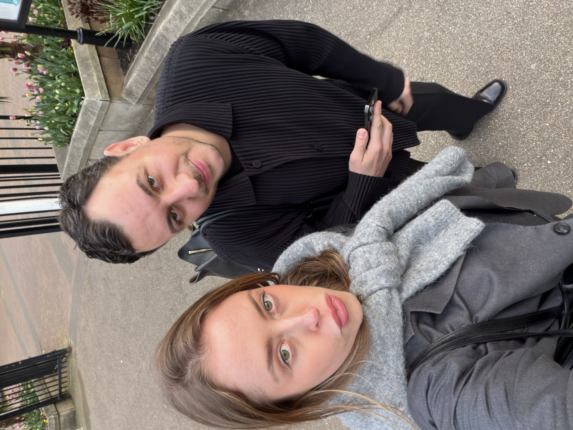 A high-angle photograph captures two individuals, a female and a male, standing on a grey paved pathway in an urban outdoor environment in London, United Kingdom. The female, positioned in the foreground on the left, has light brown hair, fair skin, and green eyes, and is wearing a grey knit scarf over a grey blazer. She looks directly at the camera with slightly puckered lips. The male, positioned to her right and slightly behind, has dark hair, stubble, and brown eyes, with a small gold stud earring in his left ear. He is wearing a black pleated collared shirt, black trousers, and black patent leather shoes, and holds a black smartphone horizontally in his right hand. He looks towards the camera with a subtle smile. In the background, behind the male, a raised concrete planter box contains blooming pink and yellow tulips, purple pansies, and green foliage, including a partial view of a palm tree. Black metal railings are visible behind the planter.