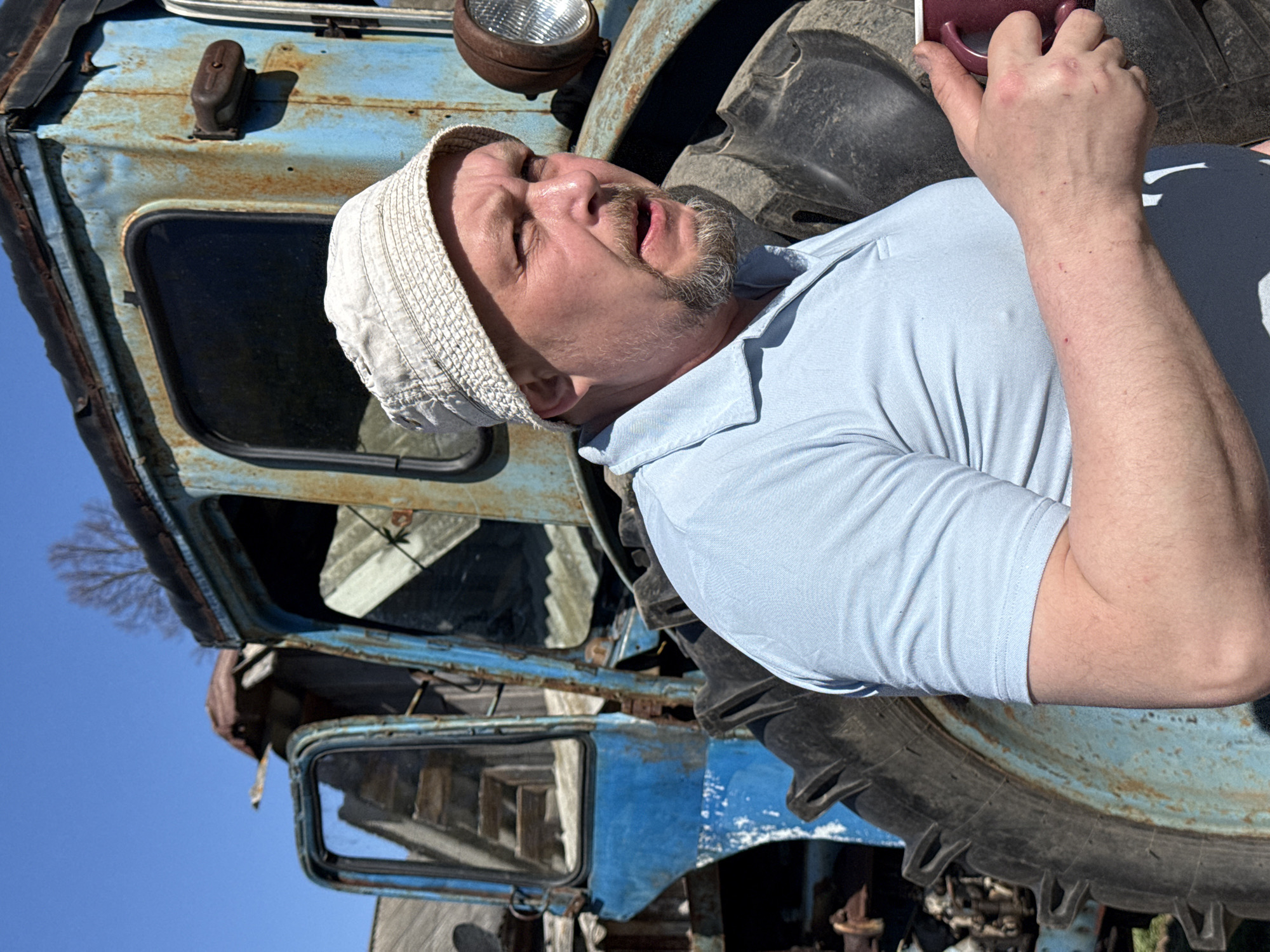 A man wearing a light-colored collared shirt and a white brimmed hat is lying beside the large tire of a weathered blue tractor. He is holding a dark-colored mug in his right hand. The tractor shows signs of rust and age. The location appears to be outdoors under a clear blue sky. Though Nizhnyaya Chermoda, Russia, is noted, there are no specific identifiable features to confirm the location.