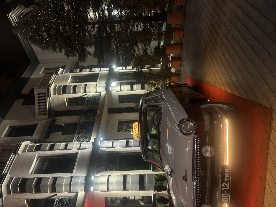 A vintage car, predominantly white with a dark roof, is parked on a red carpet. The car's front grill and a license plate are visible. Behind the car, there's an Art Deco building with a series of windows and geometric patterns. The building is lit up by spotlights. Above the building, there are trees illuminated with small lights, and a line of terracotta pots adds to the scene. The photograph is taken at night.