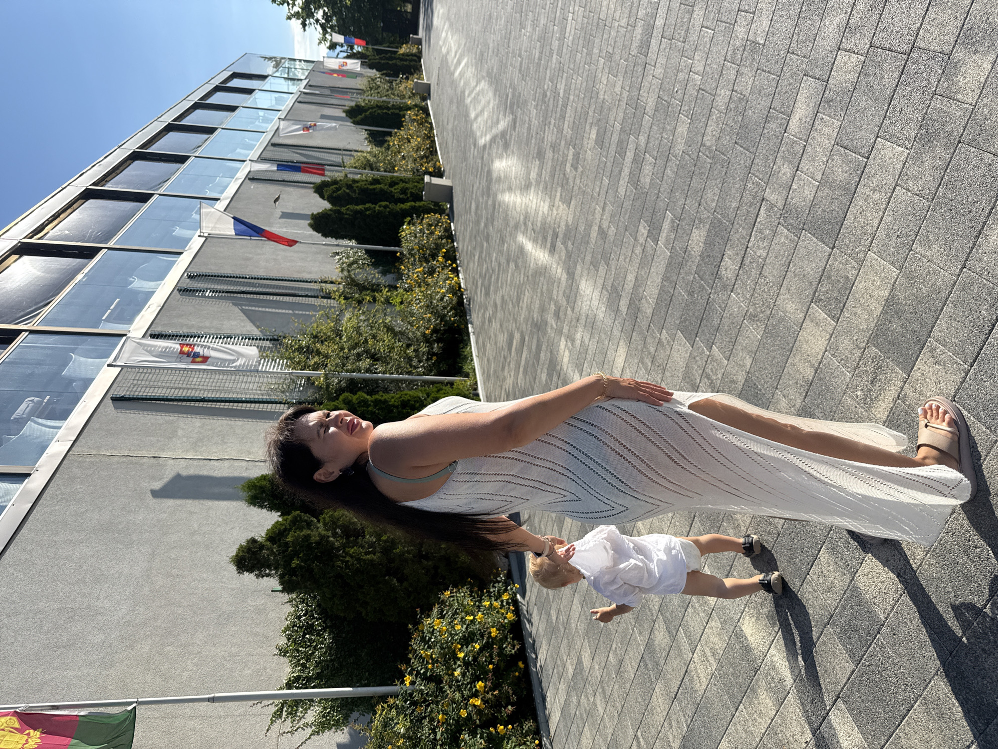 A woman and a toddler are walking on a paved sidewalk in front of a modern building with glass windows. The woman, with long dark hair, is wearing a white, sheer, sleeveless dress with a high slit and sandals. She is looking over her shoulder with a slight smile. The toddler, facing away from the camera, is wearing a white shirt and diaper, and dark shoes. They are walking towards the right side of the image. The building appears to be a hotel or resort, with flags of various countries displayed along its facade. The area is well-maintained with landscaping, including bushes and trees. The lighting suggests it is daytime, likely late afternoon, given the long shadows cast by the people and objects. The environment suggests a public space in a warm climate, consistent with Sochi, Russia.