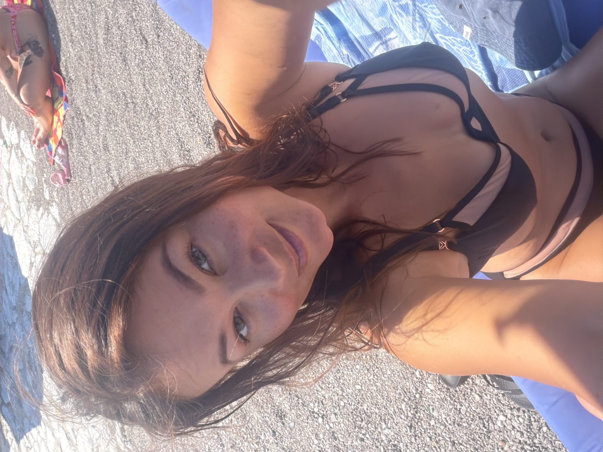 A woman in a black and pink bikini is sunbathing on a beach in Prijevor, Montenegro. She is lying on a blue towel with a grey hat next to her. The beach is covered in pebbles, and there are other people in the background also sunbathing. The woman is looking at the camera and smiling. The sun is shining brightly, casting shadows on the scene.